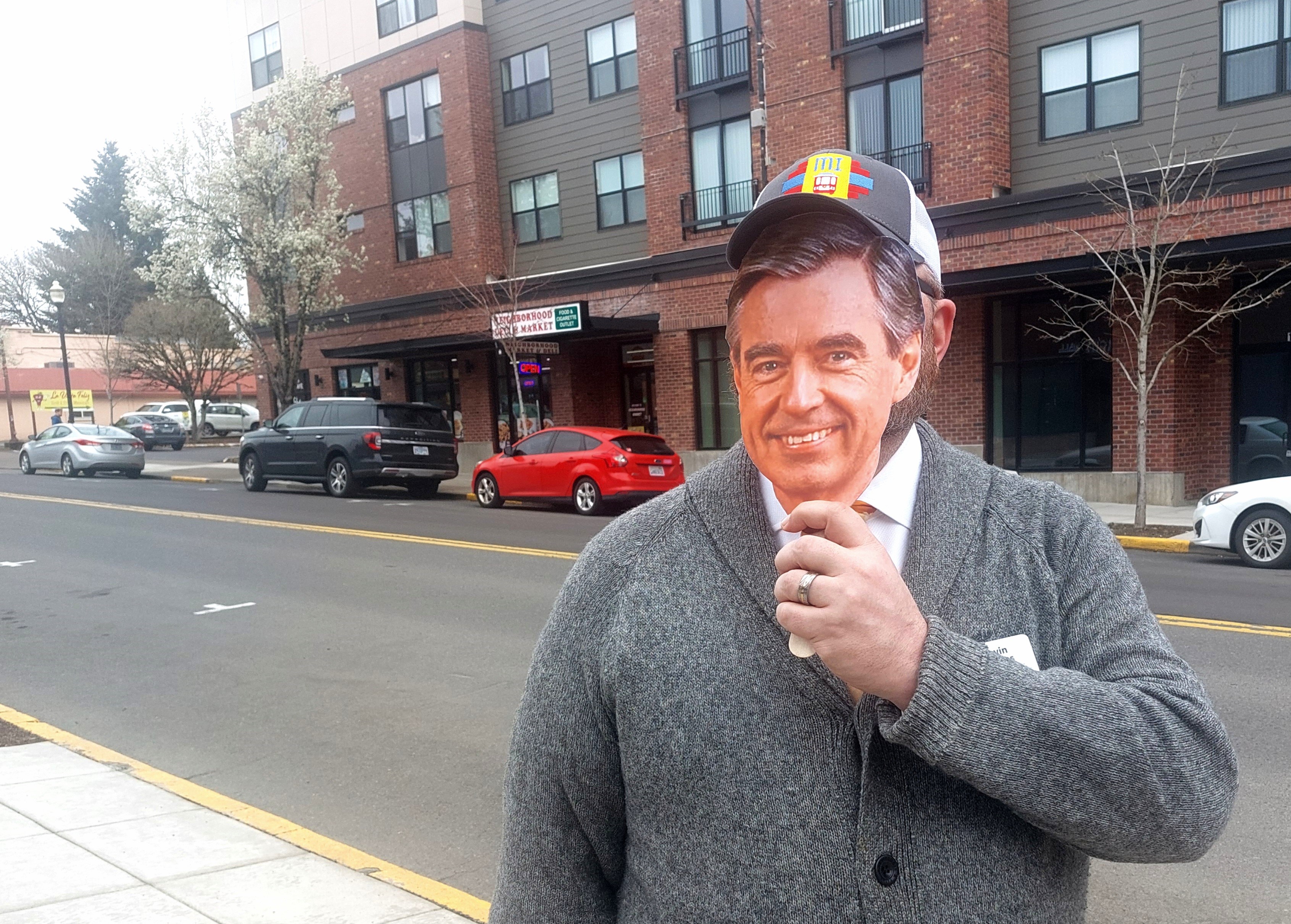 Wearing a gray cardigan sweater and holding a photo of fred rogers in front of his face, Kevin Parks poses for a photo