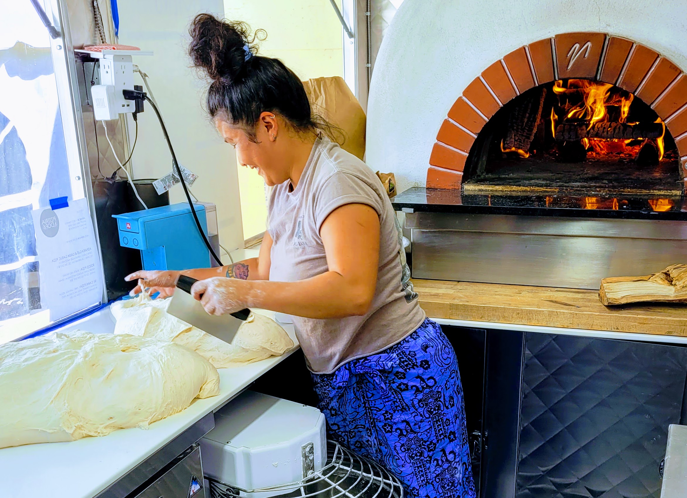 A woman cuts pieces of pizza dough from a large ball of dough. A wood-fired oven is in the background.