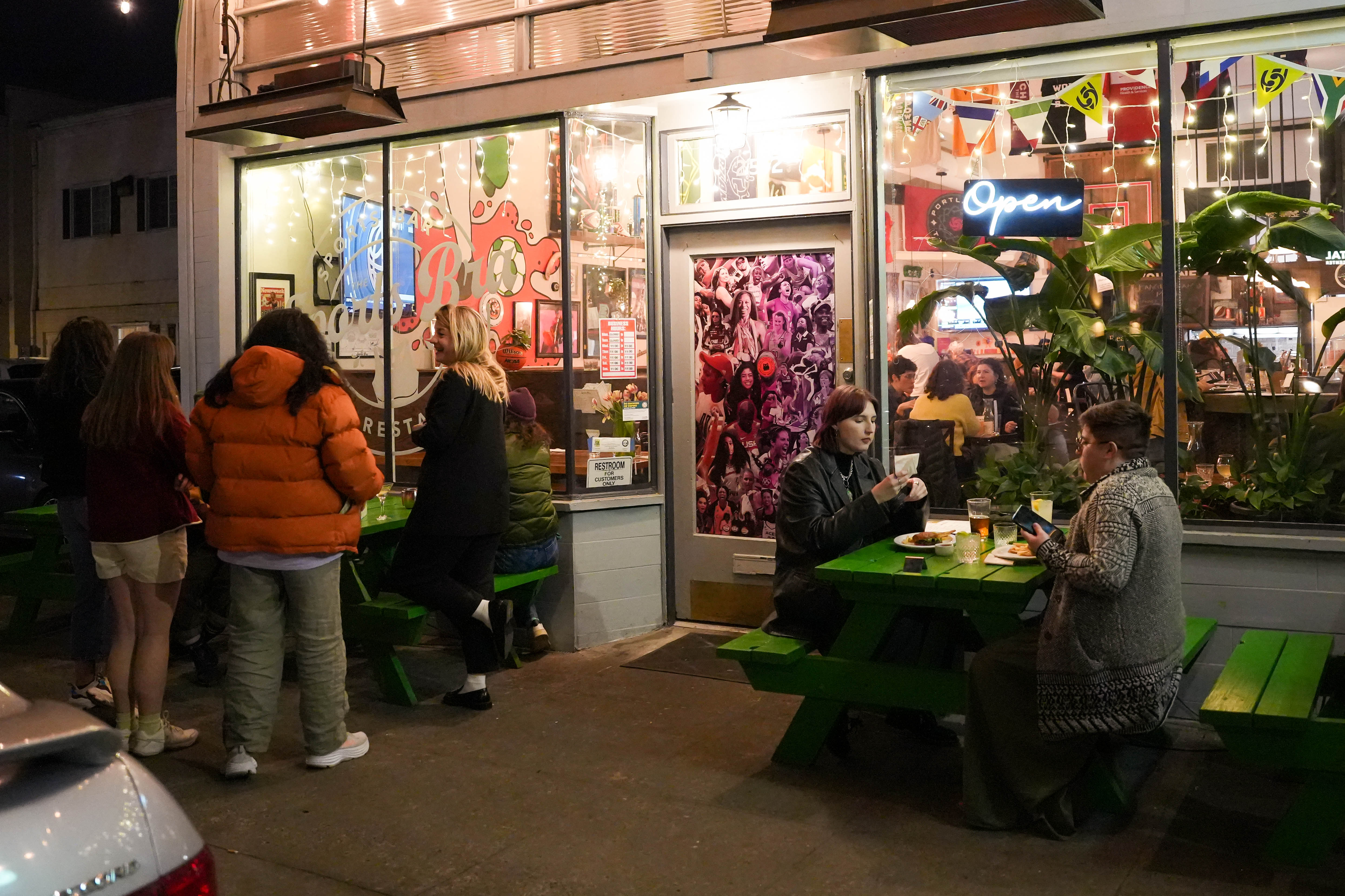 People stand outside restaurant with green picnic tables at night