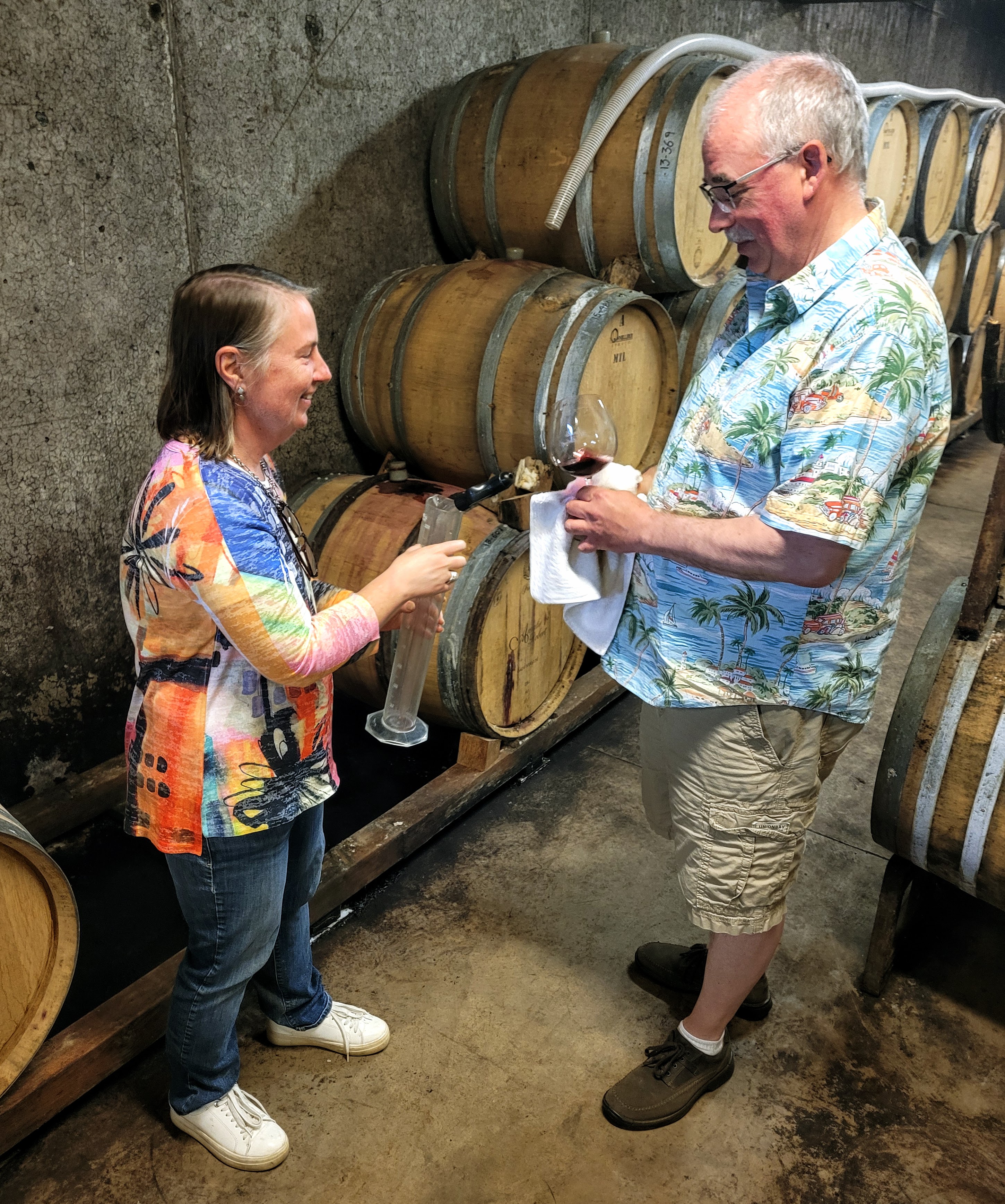 A woman and a man take a sample from a wine barrel, using a funnel-like device to transfer a sample to a beaker.