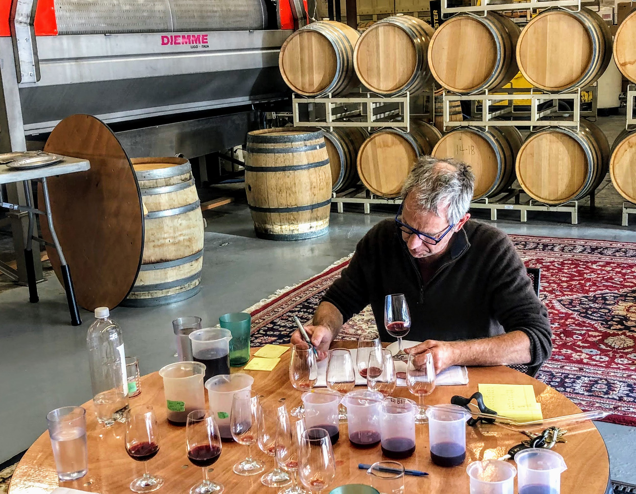 A winemaker takes notes while sitting at a table in a winery barrel room. The table is covered with wine glasses and plastic containers holding barrel samples of different red wines.