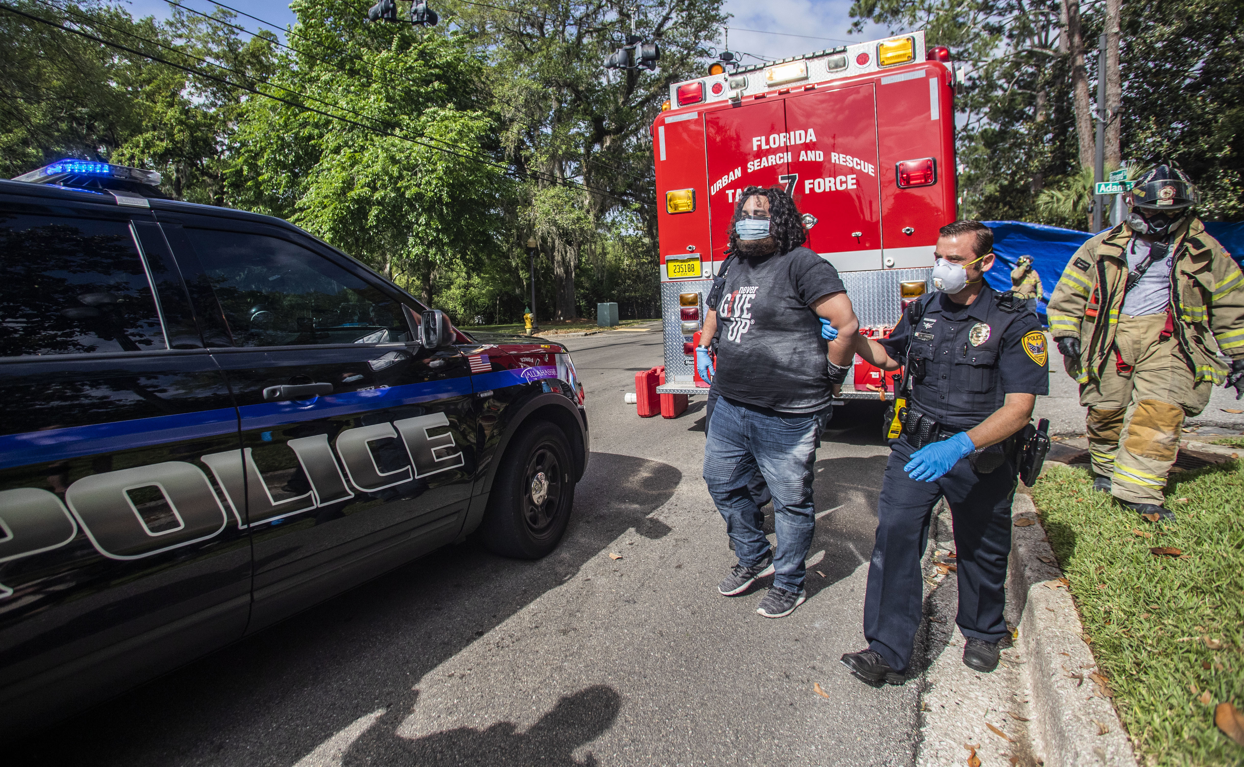 Protester Jordan Mazurek, 28, who cemented his hands in two 55-gallon plastic drums filled with concrete outside of the Governor's Mansion, is escorted by a Tallahassee Police Department officer in handcuffs to a vehicle, Friday, April 17, 2020. Mazurek is protesting how Gov. Ron DeSantis is handling coronavirus in state prisons. (Alicia Devine/Tallahassee Democrat via AP)