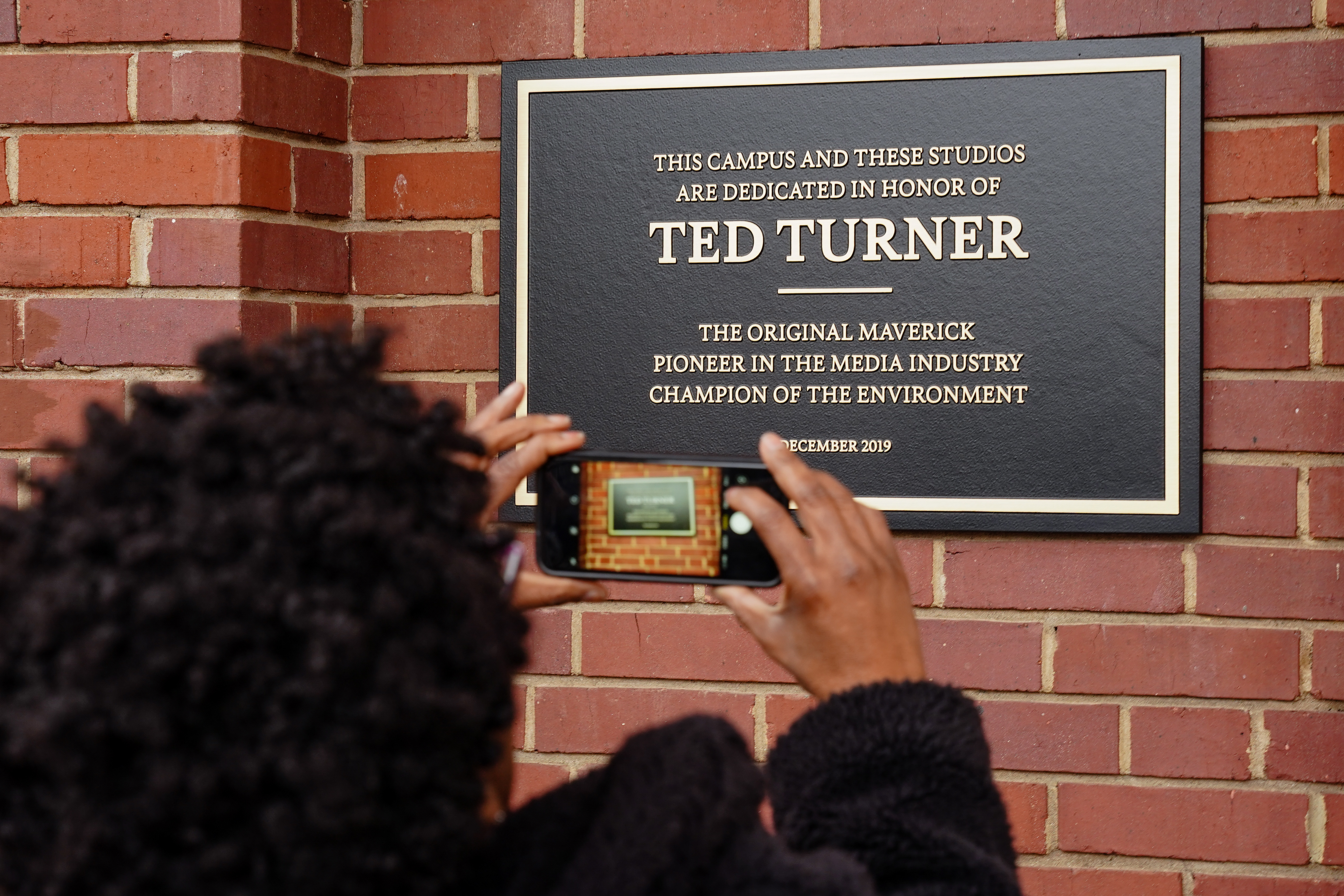 A woman takes a photo of a plaque on an AT&T WarnerMedia building dedicating it to Ted Turner, the founder of Turner Broadcasting System, on Friday, December 6, 2019, in Atlanta. (Elijah Nouvelage/Special to the Atlanta Journal-Constitution)