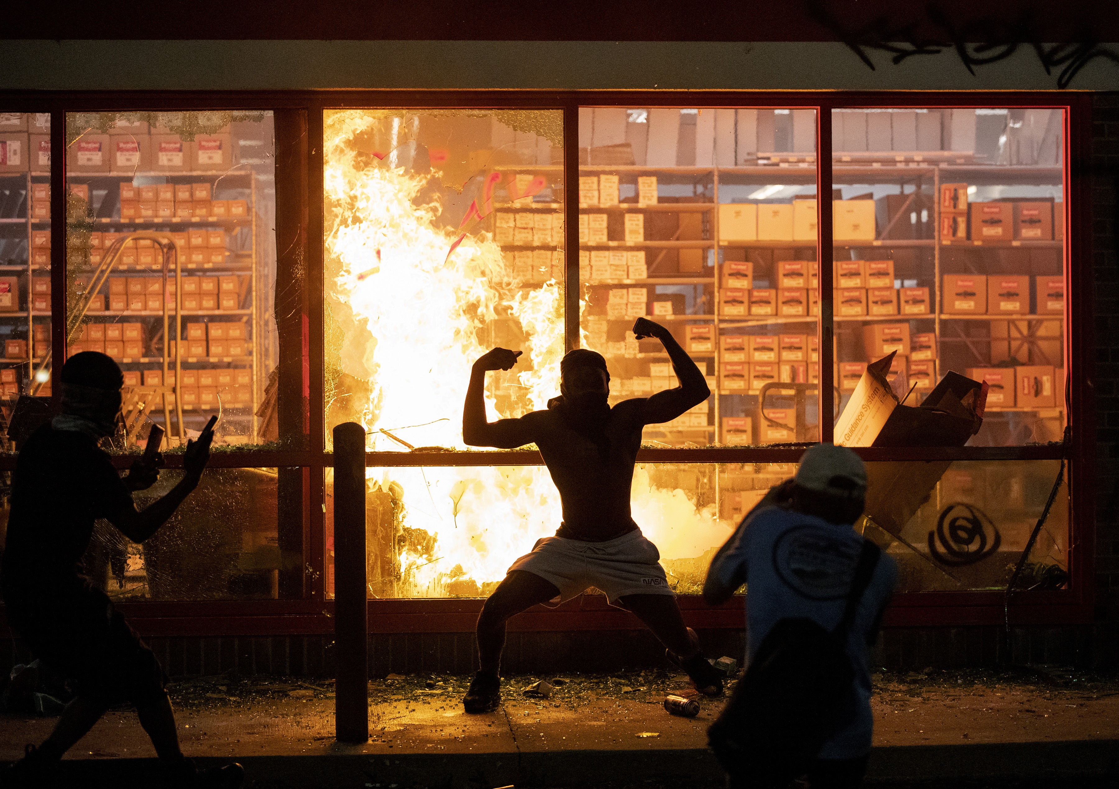 A man poses in front of a fire at an AutoZone store, while protesters hold a rally for George Floyd in Minneapolis on Wednesday night.
