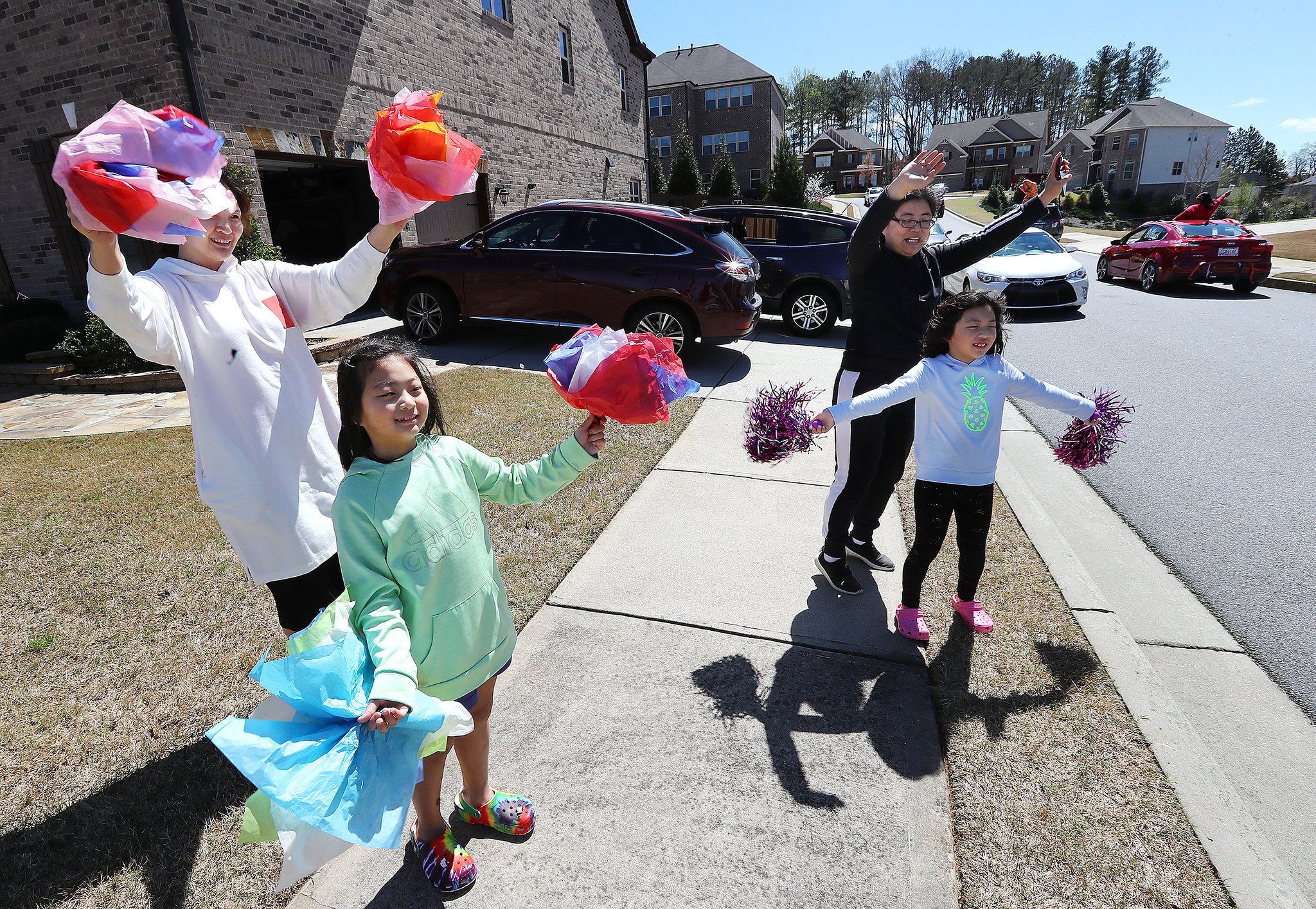 Lydia Suh (from left), her daughter Rachel and Nancy Acosta, and her daughter Mei use tissue paper and pom poms to cheer their teachers from Roberts Elementary as the teachers drive through nearly two dozen area neighborhoods for some face-to-face contact while still maintaining social distance to ease the separation anxiety of their students during the coronavirus on Wednesday, March 25, 2020, in Suwanee. Although distance learning is allowing kids to connect with teachers and get lessons done, the personal touch is missing. CURTIS COMPTON CCOMPTON@AJC.COM
