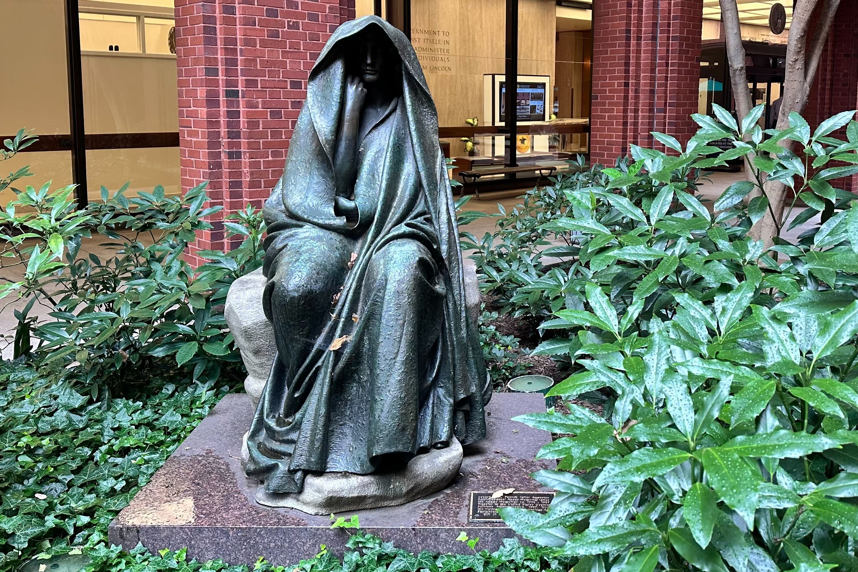 The "Black Aggie" statue sits in a courtyard in Washington, D.C. The statue depicts a hooded figure with its chin resting on its fist in a contemplative stance. The statue is surrounded by green foliage.