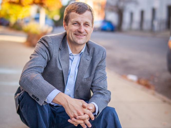 A photo of Mark Parker sitting on steps wearing a gray suit jacket, a light blue shirt, dark blue slacks, and brown shoes.