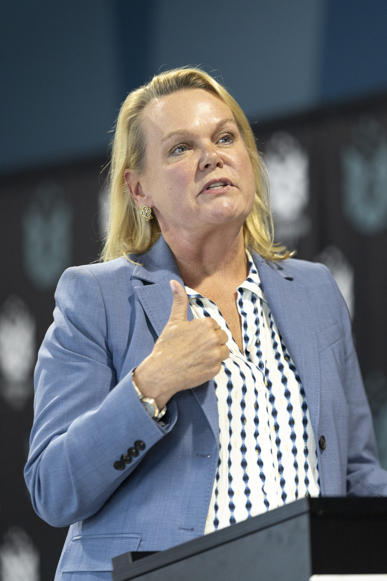 April McClain Delaney is wearing a blue suit jacket and white shirt with blue dots while speaking at a lectern.