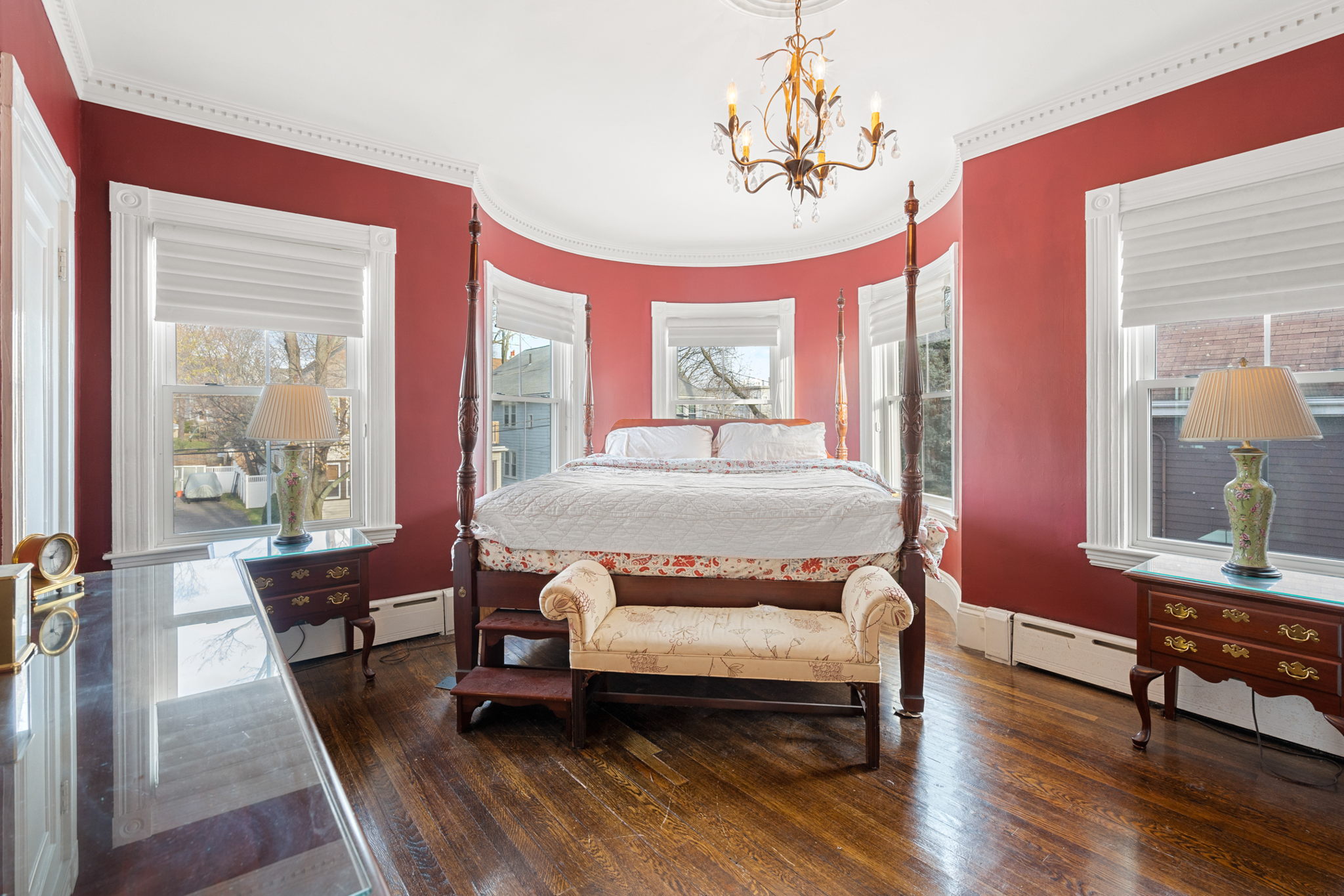 The owner suite has red walls, a brass and crystal chandelier, hardwood flooring, crown molding. the trim is white. There is a two poster bed with a white duvet, a settee in front of the bed, a small bureau with a shaded lamp, an end table with a shaded lamp, and a bureau.