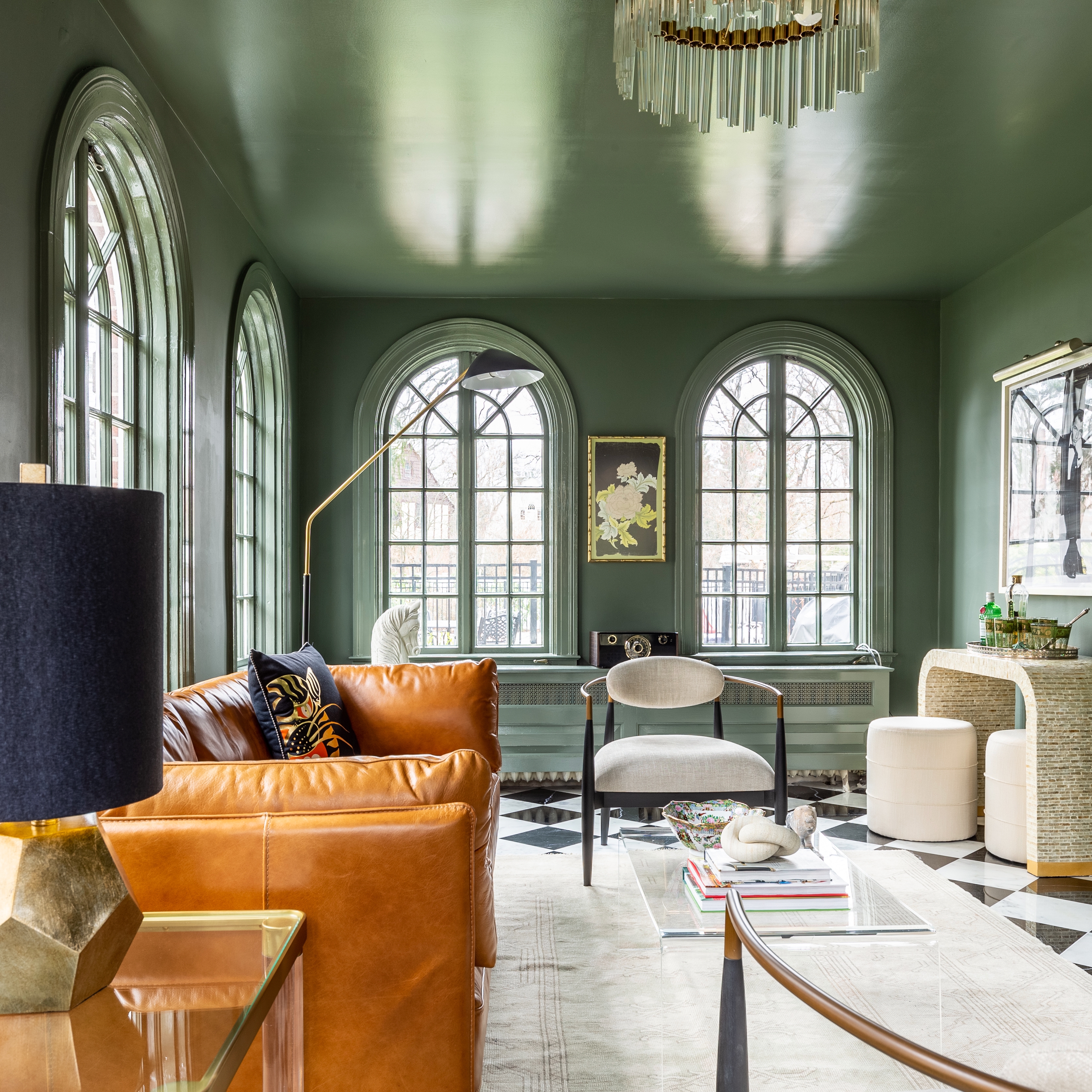 The ceiling and walls in this room are painted a dark sage green and so are the window frames, as well as the radiator that lines the back wall. The windows are tall, wide, and arched. To the left is a bright brown leather couch with a black throw pillow, a glass table with gold framing, and a bulbous, geometric lamp with a black shade. In the middle of the room in the foreground sits a glass, gold-framed coffee table. In the back middle is a wide armchair with an off-white cushion. To the back right is a modern, arched bar with two poofs for seats. Bar implements sit on top.
