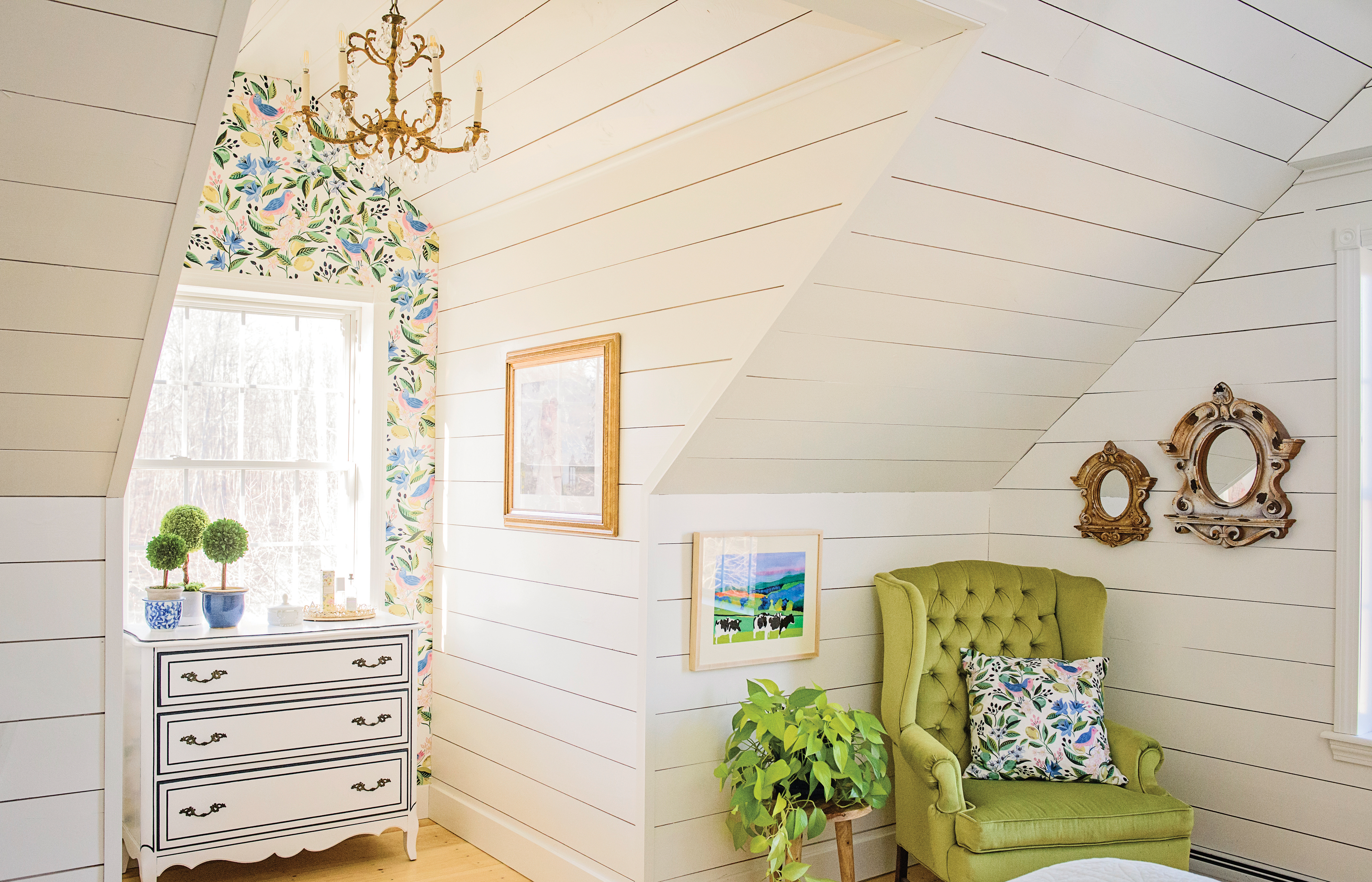 The dormer of the primary bedroom has Caitlin Wilson wallpaper, a gilded chandelier from The Collector’s Eye in Stratham, New Hampshire, and a dresser that homeowner Amber Roy refurbished. “I take any old furniture offered to me,” she says. The cow painting by Woody Jackson is from Edgewater Gallery in Middlebury, Vermont.