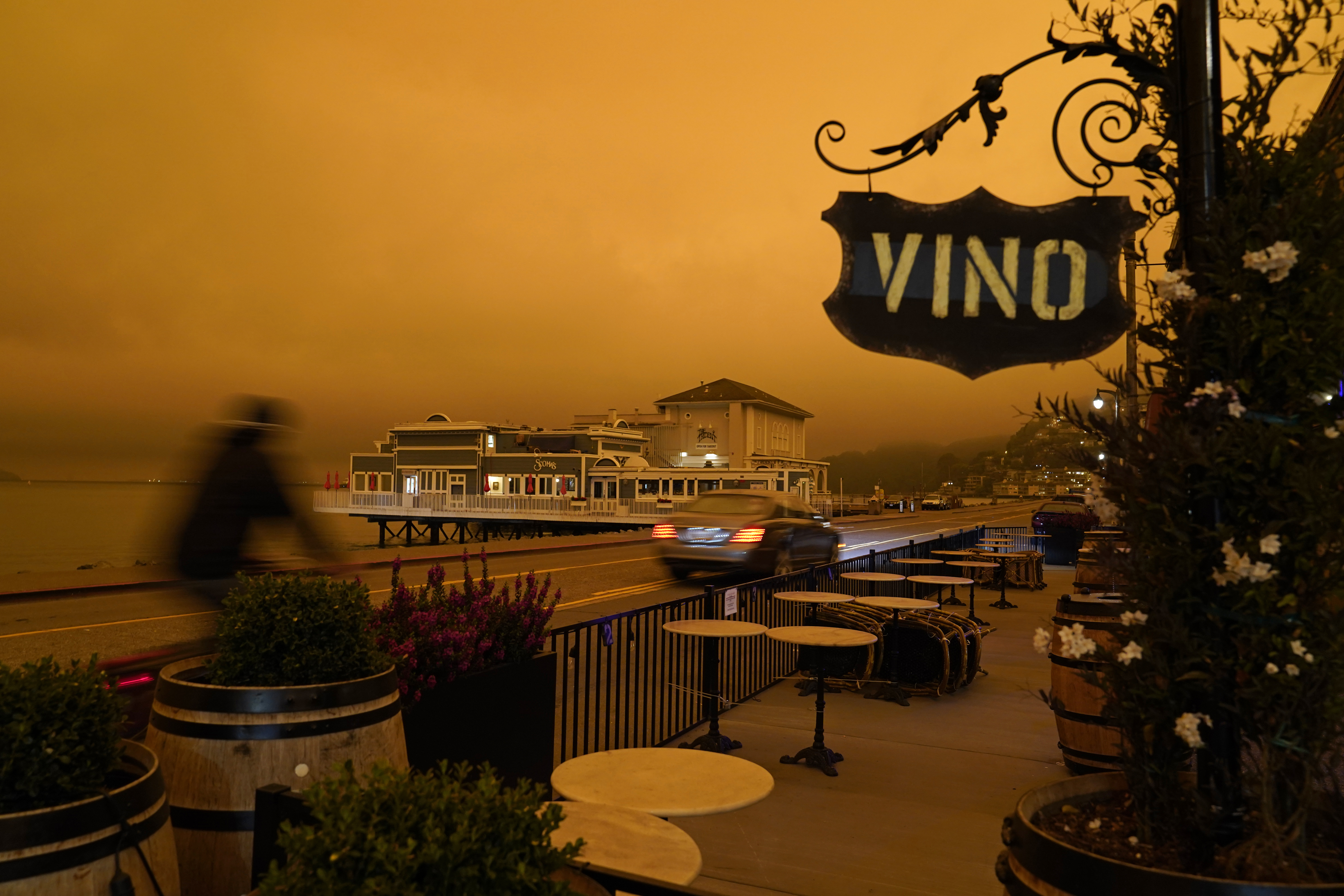 A bicyclist rides past a seating area outside a wine bar as smoke from wildfires darken the morning sky, Wednesday, in Sausalito, Calif. (AP Photo/Eric Risberg)