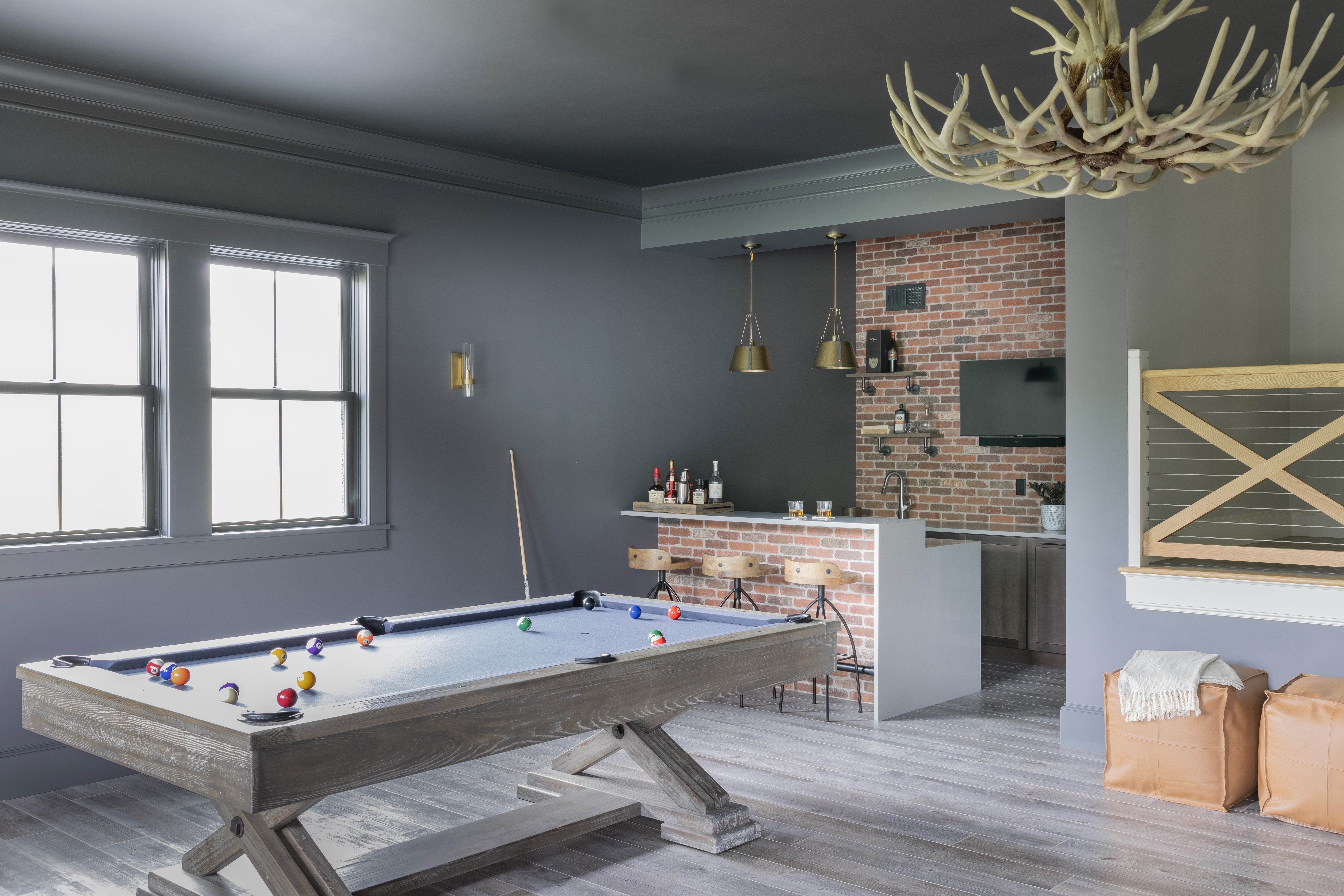 A driftwood-colored pool table sits before a pair of windows. There are two orange ottomans to the right and a wooden stair rail in the shape of an X. To the left, tucked in a corner is a brick bar with bottles on top and three round stools in front. Two pendant lights with a bronze look hang above. The brick ack wall has a mounted counter, open shelving, and a TV.