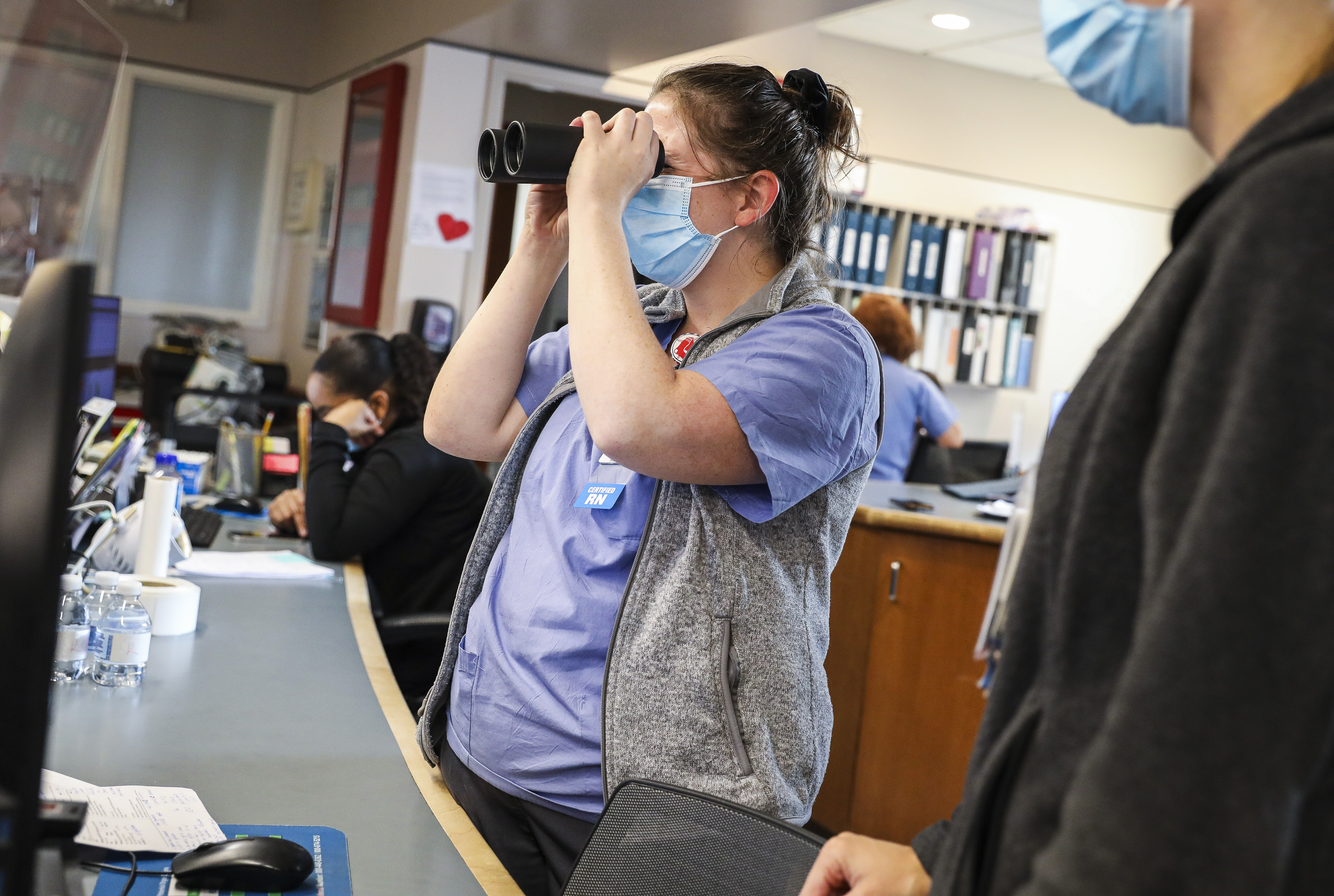 Tufts Medical Center nurse Katey Ring used a pair of binoculars to look through the glass walls of a patient's room while working in the medical intensive care unit.  To limit exposure to COVID-19 and preserve PPE, binoculars were used during the pandemic to assess patient pumps and ventilation settings on the ground. 