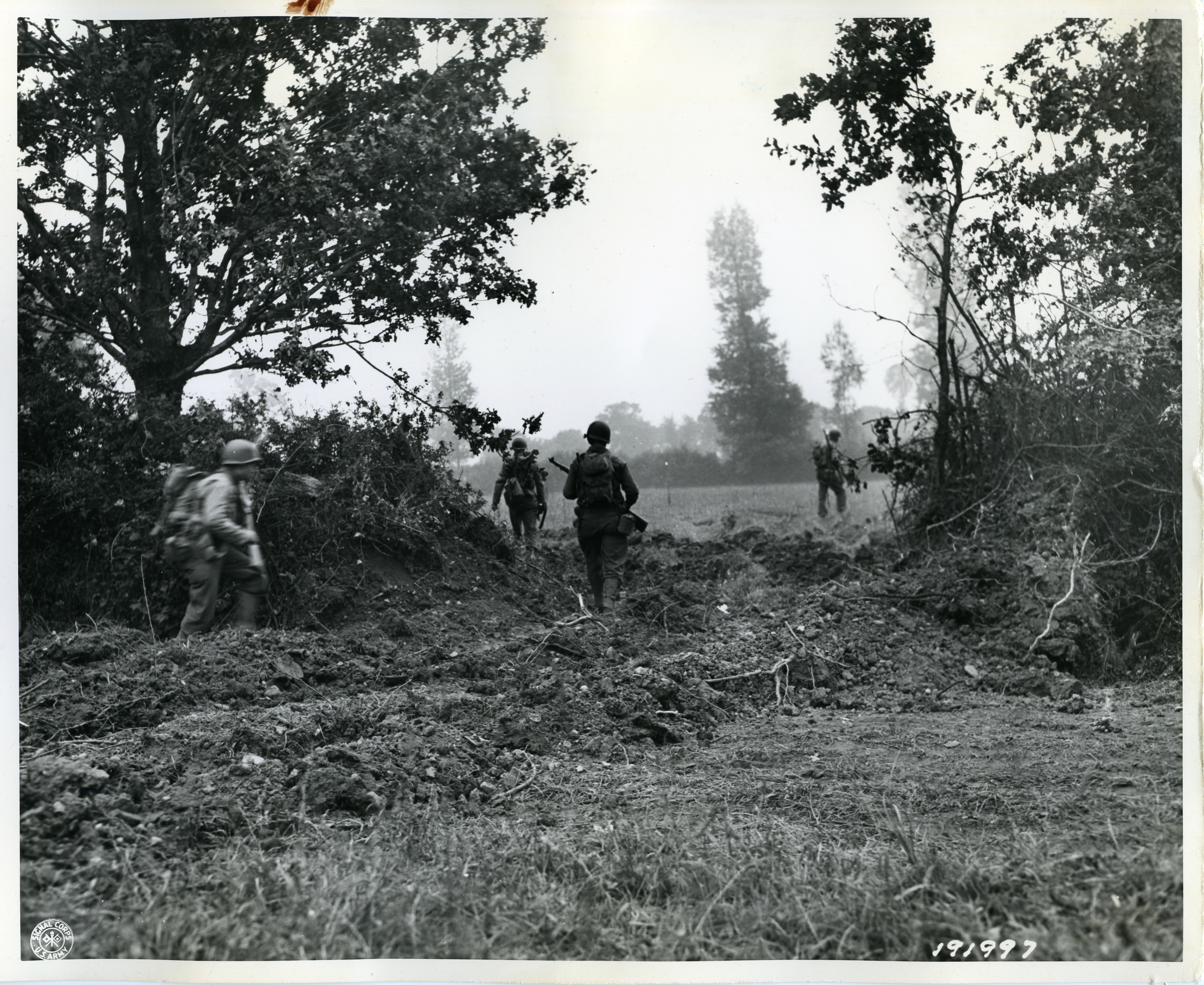 GI's move forward through a breach in a hedgerow made by a bulldozer during World War II.
