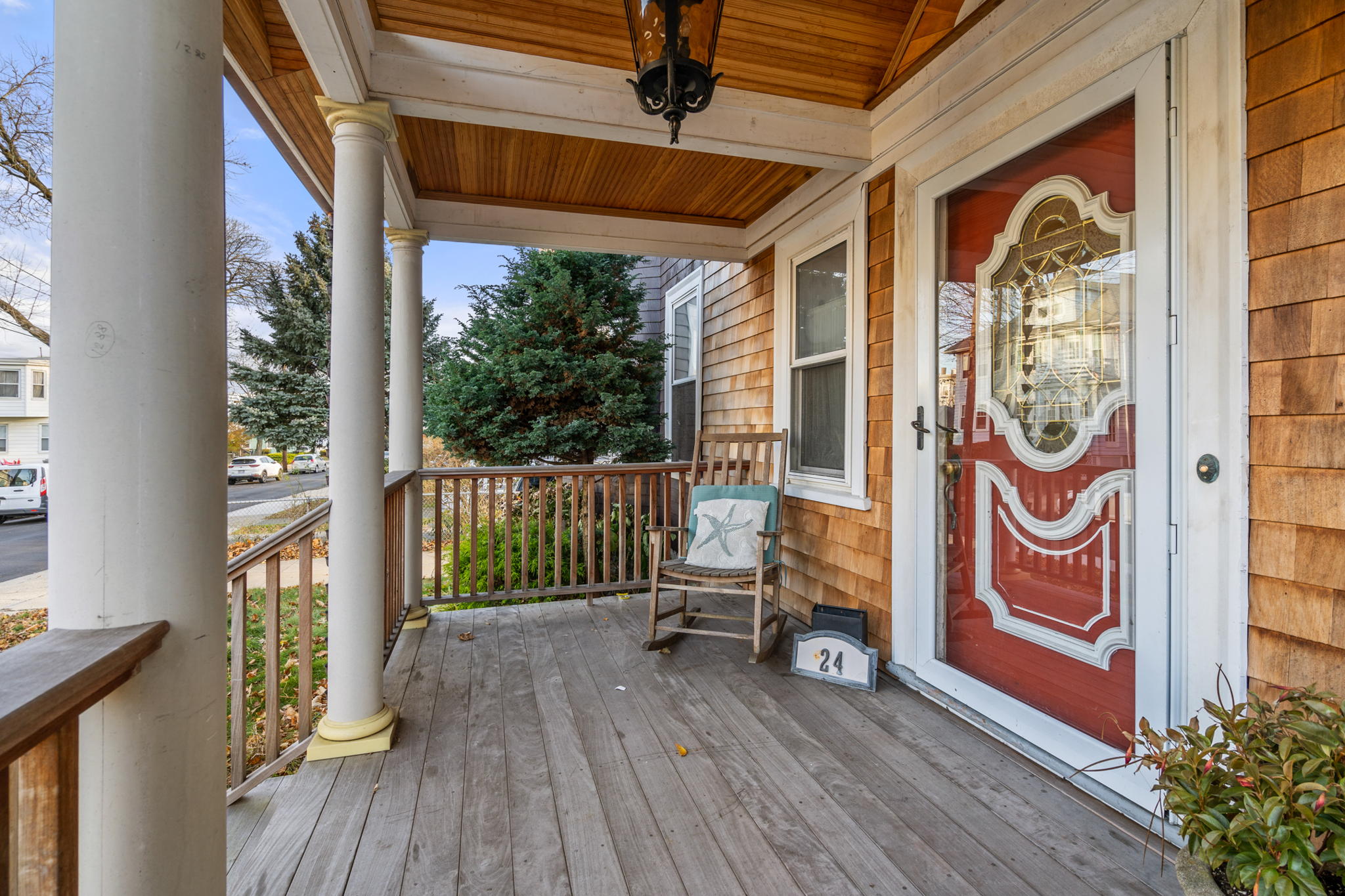 A view of the home's wooden front porch, which has white columns, cedar shake shingles, a black lantern, a rocking chair, a blue box on the planks with the house address number on it (24), and a red front door with elaborate white trim that looks almost Moroccan.