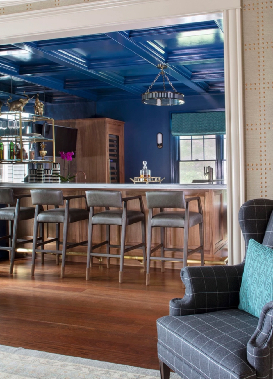 The coffered ceiling and the walls in this space are painted a moody dark blue. There is a wing chair in the foreground with a blue checkered print and a teal, patterned pillow. The bar is in the back on step up. It has a white counter, four hard-backed metal stools, and a brass foot rail. There is a window behind the bar, and the sunlight is bouncing off the glass backsplash behind open shelving filled with liquor bottles. There is a carafe on top of the bar.