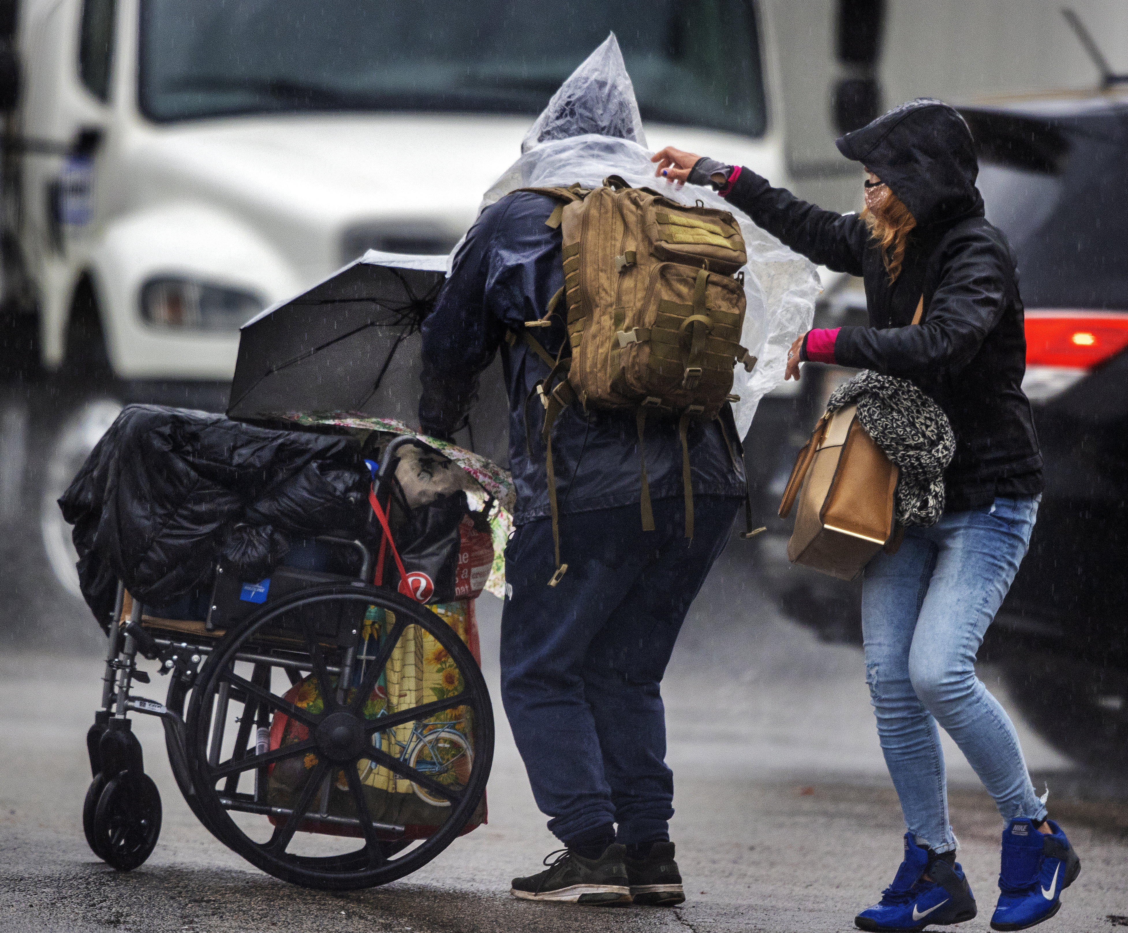 A homeless man pushes his belongings in a wheelchair as a woman helps him put on a plastic poncho.