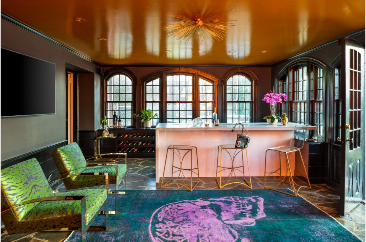 A bar with three stools sits in the background of a room with arched windows. There is a blue rug with a bright pink, bean-shaped design in the middle. Off to the left are green, zebra-striped chairs. The walls are white, as is the bar. There are pink flowers, a liquor bottle, and two quarter-full glasses on top of the bar.