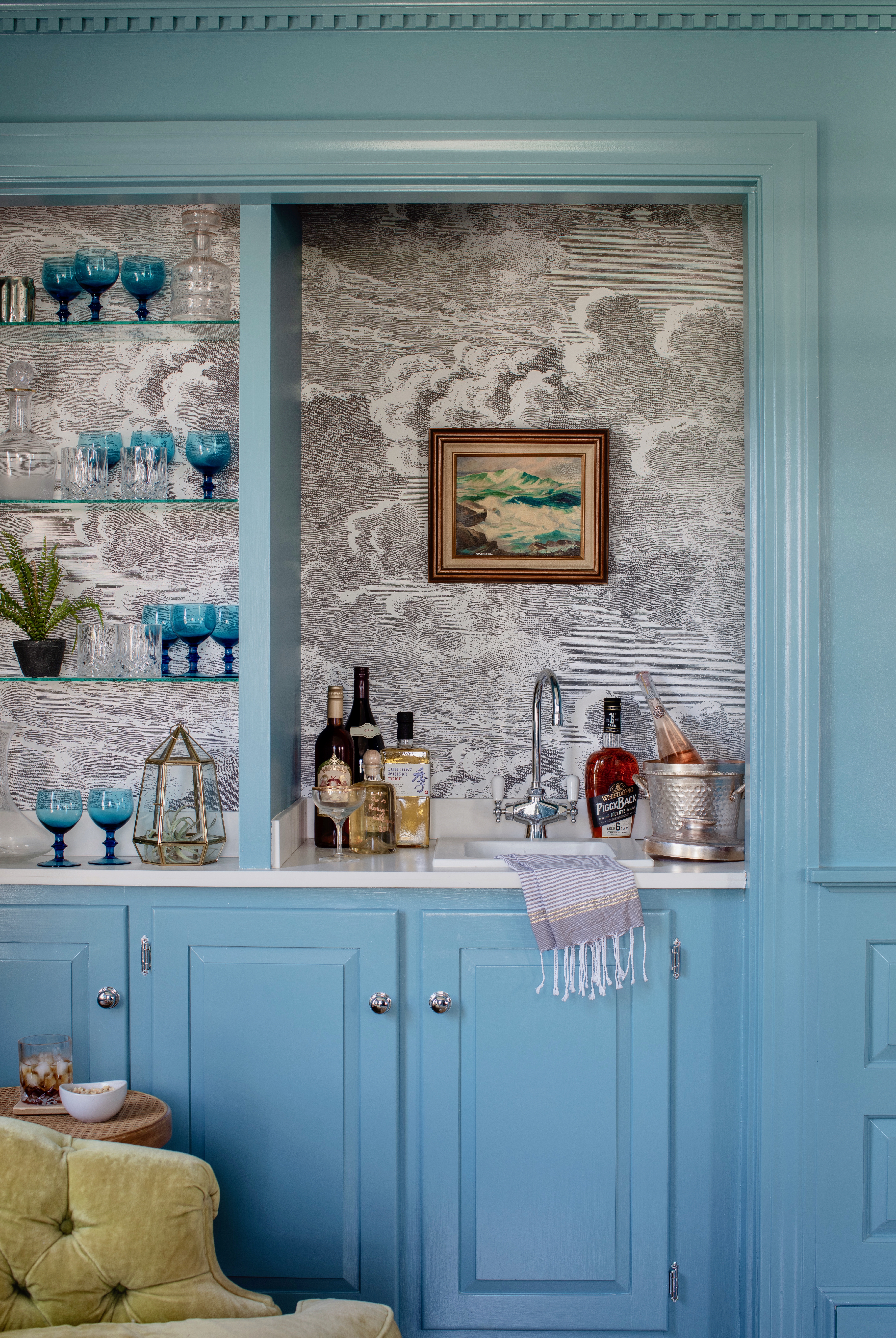 Light blue cabinetry makes up the bar in this space and matches the wainscoting and framing of the room. A fringed tea towel sits in a small sink with a metal faucet. An olive green arm chair is in the foreground next to a small round end table with a bowl and a full glass with ice on it. There are blue glasses on the glass open shelves of the bar. The backsplash is a wallpaper with a gray cloud print. There are bottles of liquor and an ice bucket with a bottle in it on the counter.