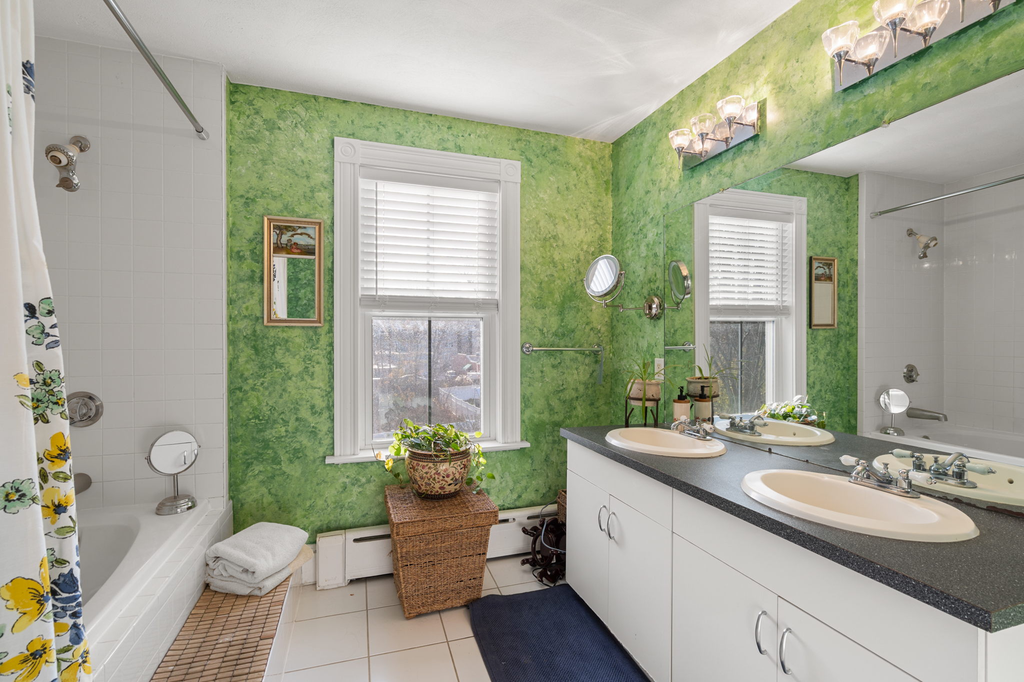 The shared bath has green walls that look like foliage, a white ceramic tile floor, a tub/shower combination behind a sunflower curtain, two sets of bulb lights above a frameless mirror, a white dual vanity with two sinks and a granite countertop, beige and navy throw rugs, and a hamper with a plant on top.