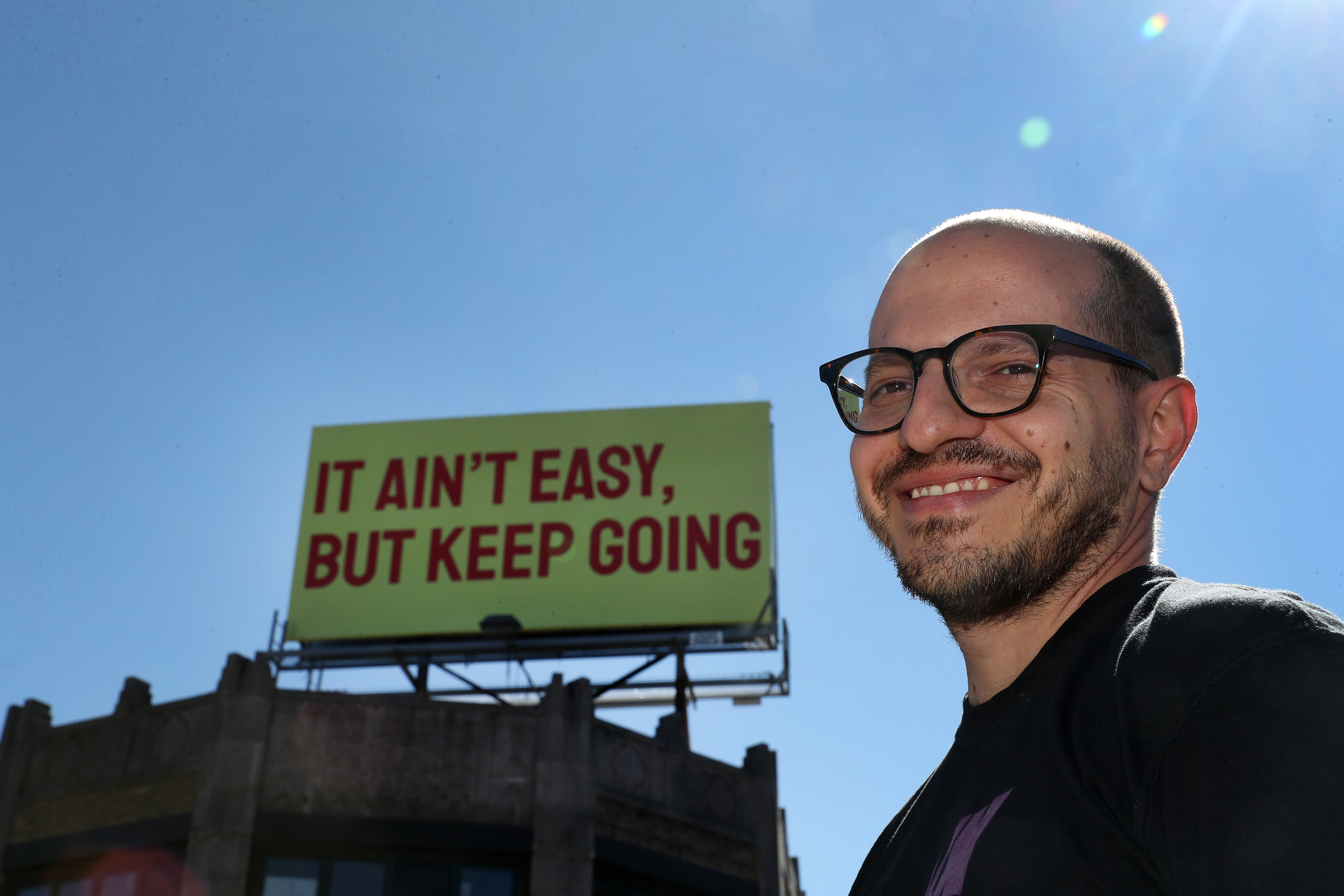 Gabriel Sosa with his "It Ain't Easy" billboard in Roslindale. Sosa has several more uplifting pieces, printed with messages in English and Spanish, soon to be installed around the city.