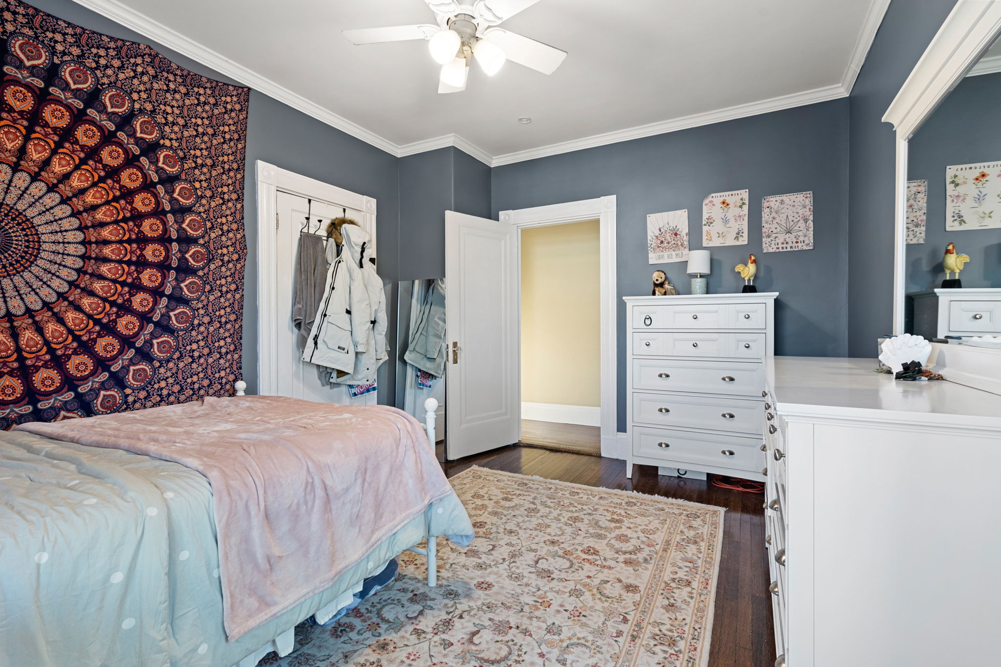 A blue bedroom with a bohemian-style fabric on the wall, blue walls, white crown molding, a bed with a pink throw, white furniture, and a ceiling fan.