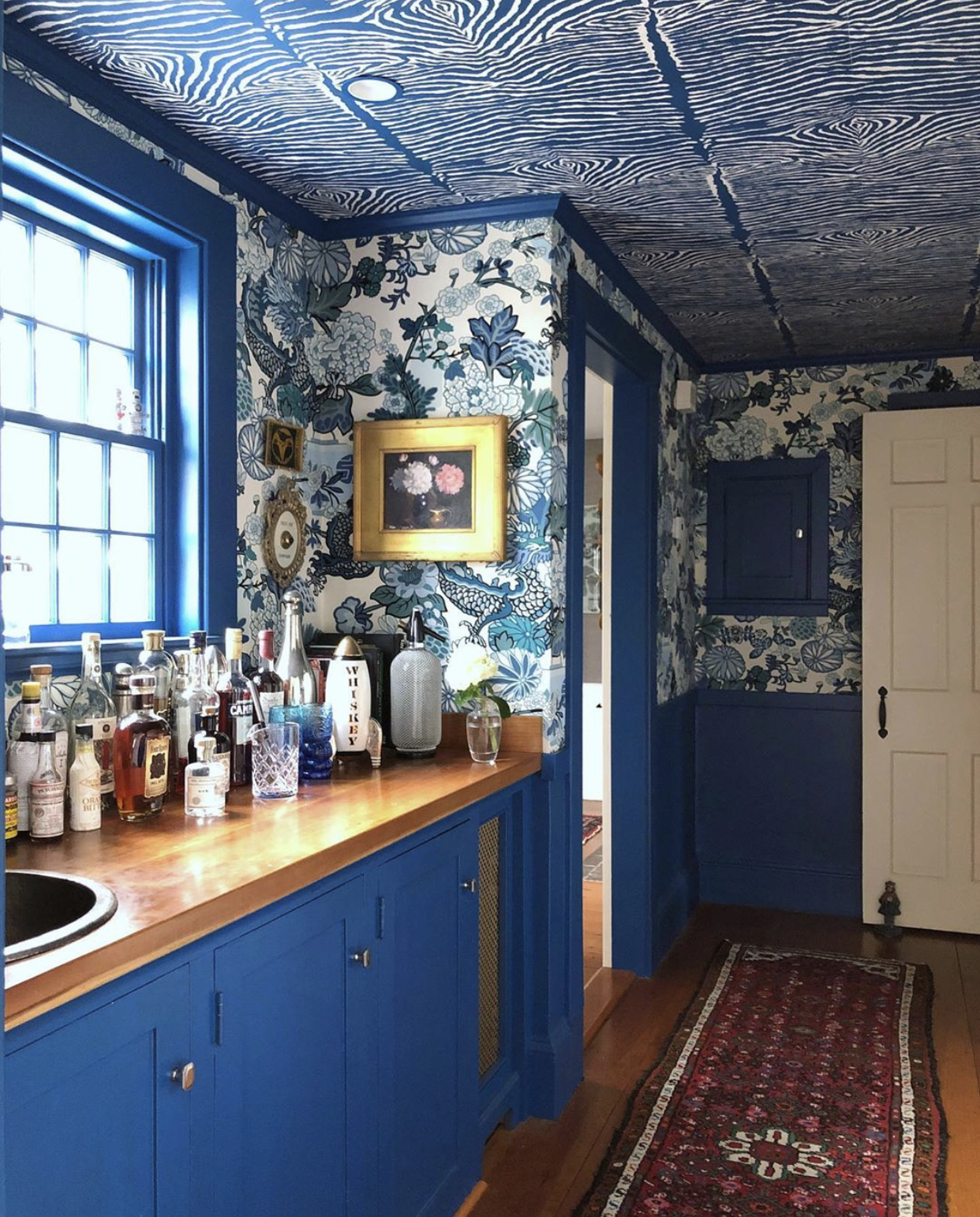 A polyurethaned counter sits atop bright blue cabinetry. Several bottles of liquor sit on top of the counter, glinting in the sunshine spilling in from the blue-framed window above. There is a red, patterned rectangular rug on the floor. The walls are paper in a big-patterned floral paper.
