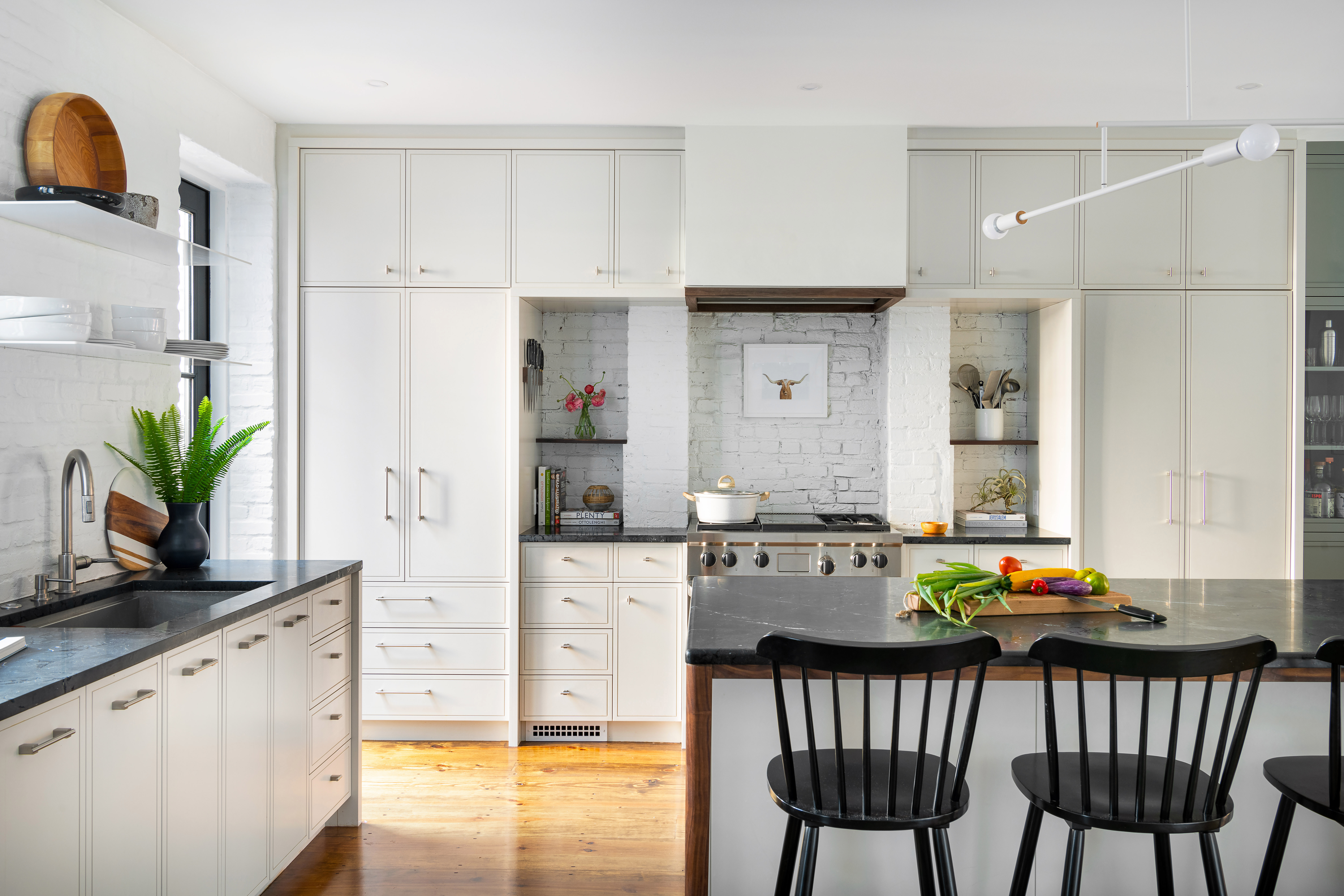Walnut details, including the shelves flanking the range and the hood trim, tie to the island. White metal shelves over the sink add a contemporary touch.
The cabinetry, made by SHAKE Architecture: Construction, is painted with Benjamin Moore Wind’s Breath to subtly contrast the bright white brick.