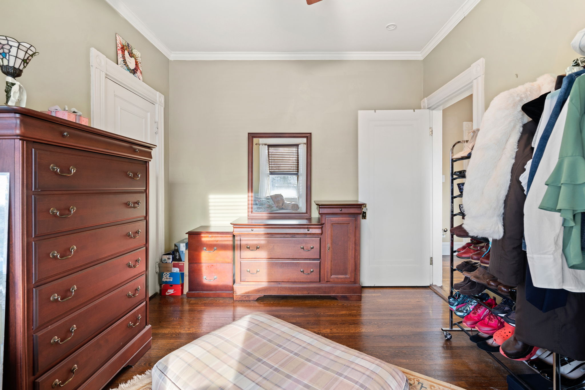 A bedroom with hardwood flooring, beige walls, white crown molding, handing clothes, shoes on a rack, wood furniture, and a beige-colored plaid rug.