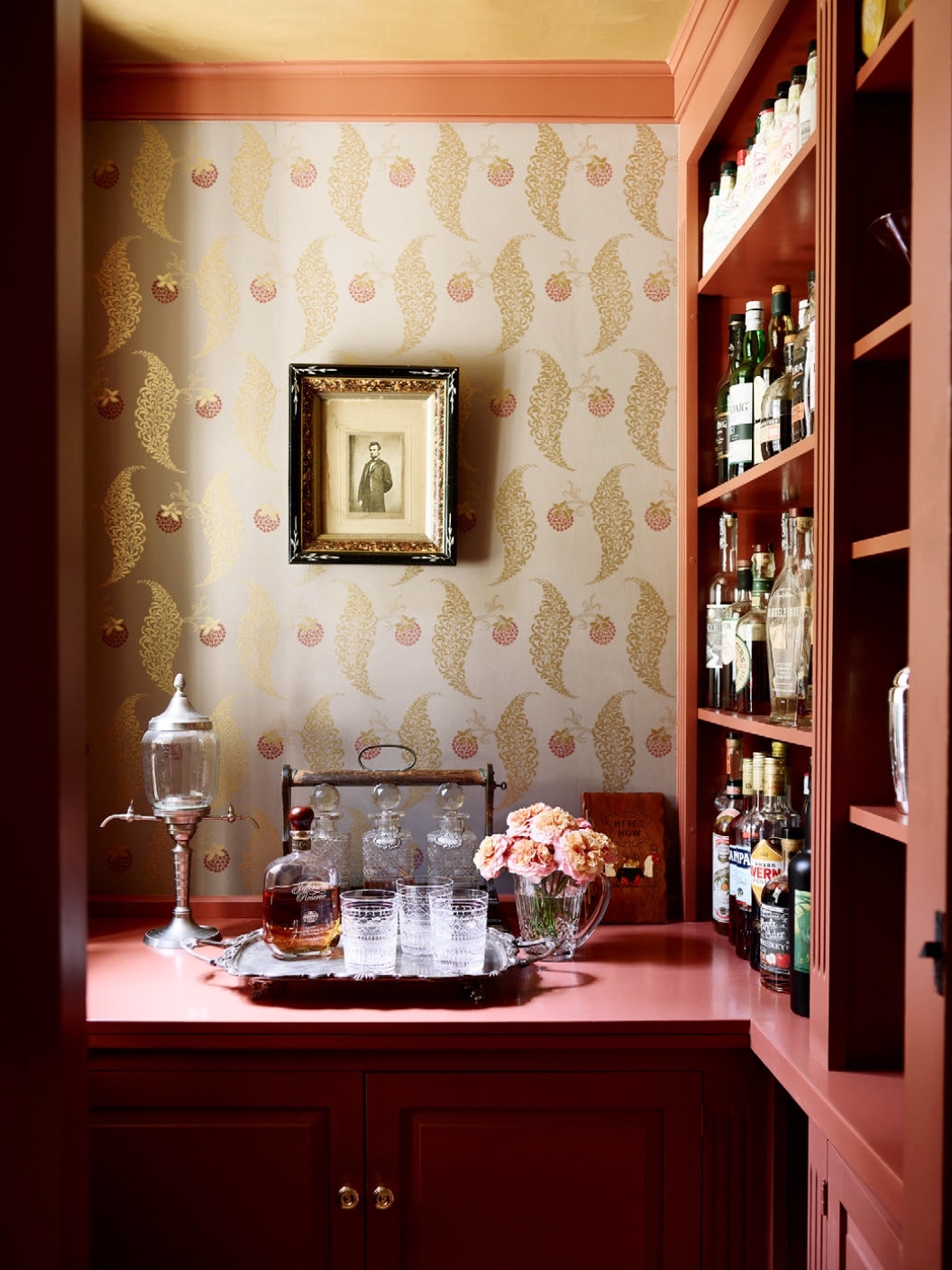 Red bar and cabinetry. The red counter has a silver tray with bar glasses and round bottle of liquor. There is a short vase of pink flowers. A lantern on a stand is to the left. The wallpaper is gold leaves and red berries. The side shelves are full of liquor bottles.