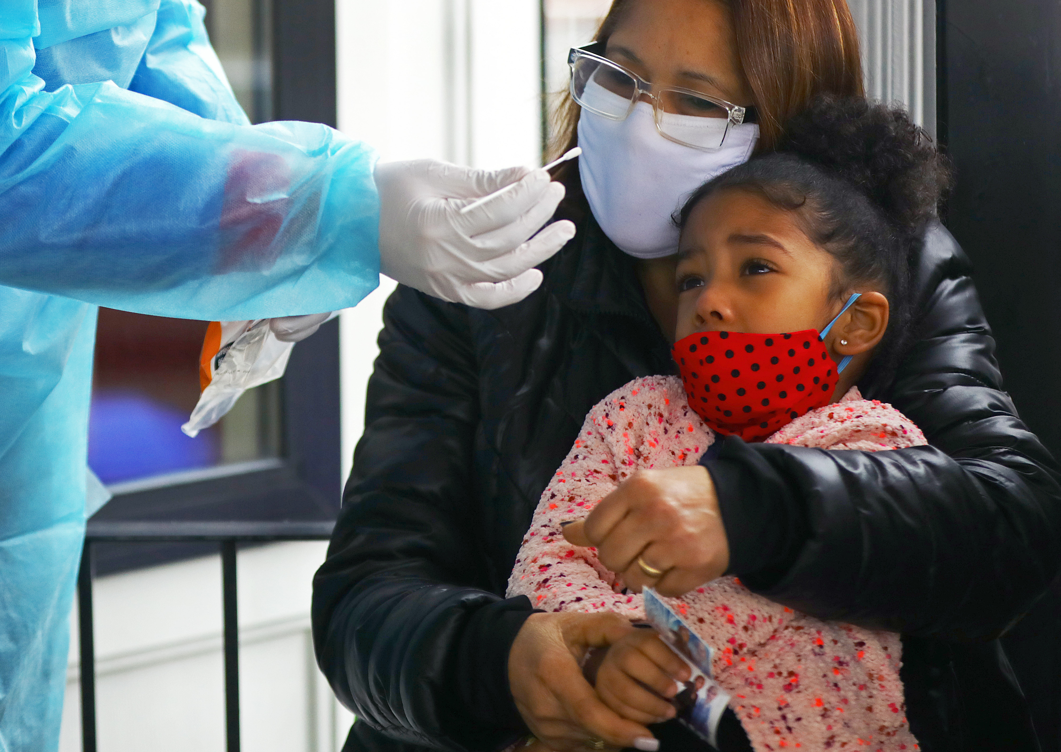 Dr. Jose Navarro attempts to get the cooperation of 3-year-old Heidy Martinez Pincel in administering a rapid antigen test and a PCR test, on the front porch of Jenks Park Pediatrics in Central Falls. Her grandmother, Nereydis Galvez, holds her through the struggle.