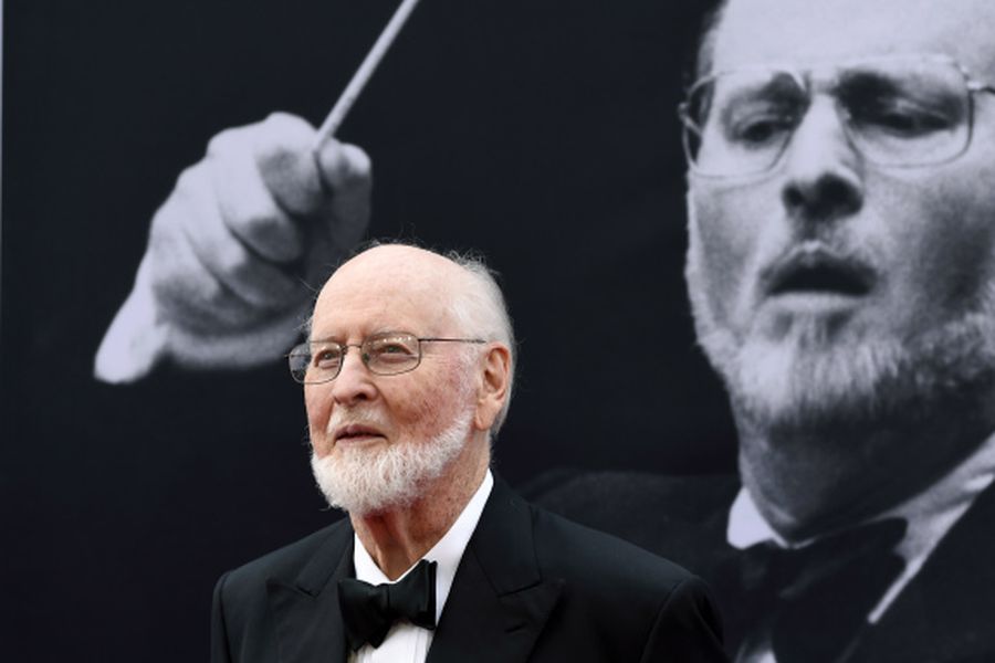 Fotografía del compositor John Williams mirando de frente. Tras de él, hay una imagen suya en blanco y negro dirigiendo con una barilla.