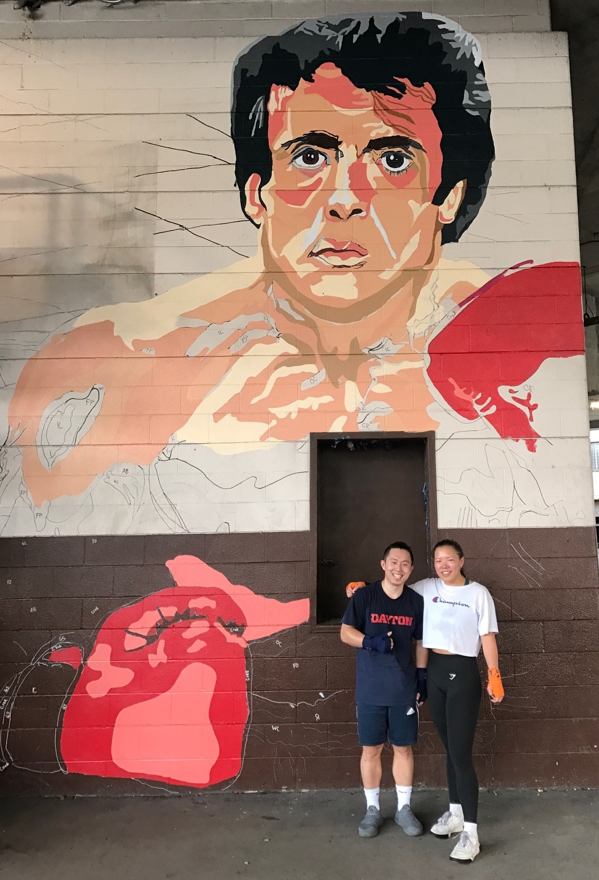 Hyde Li and Grace Ashworth in front of the large Rocky Balboa and Apollo Creed mural being painted on the side of Drake's Downtown Gym by artists from the nearby K12 Gallery & TEJAS on Jefferson Street.  Tom Archdeacon/STAFF