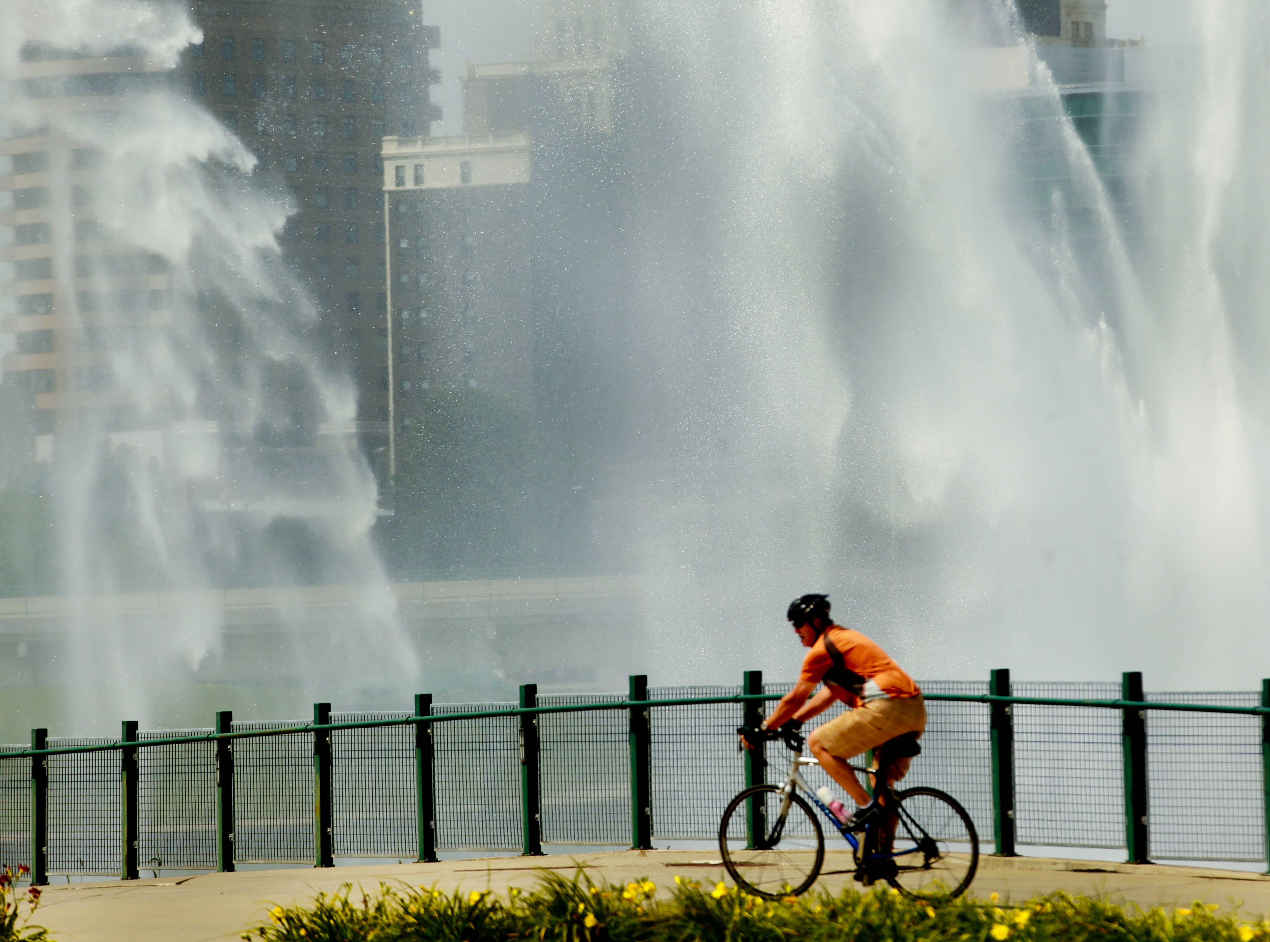 A cyclist rides along a backdrop of a noon-hour display of the spraying fountains at the Deeds Point area of Riverscape in downtown Dayton. JIM WITMER / STAFF