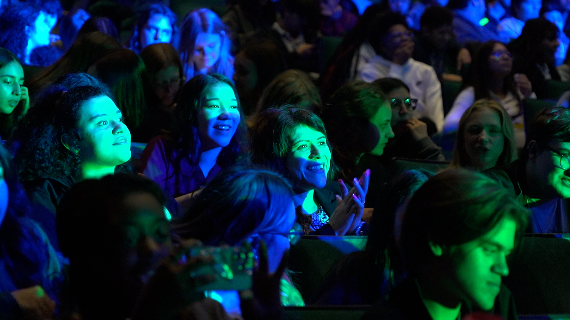 Blue and green lights shine on the faces of high school students enjoying a theater production.