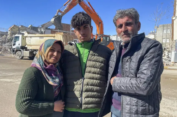 Taha Erdem, de 17 años, en el centro, su madre Zeliha Erdem, a la izquierda, y su padre Ali Erdem posan para una fotografía junto al edificio destruido donde Tahan quedó atrapado tras el terremoto, en Adiyaman, Turquía. (AP Photo/Mehmet Mucahit Ceylan)