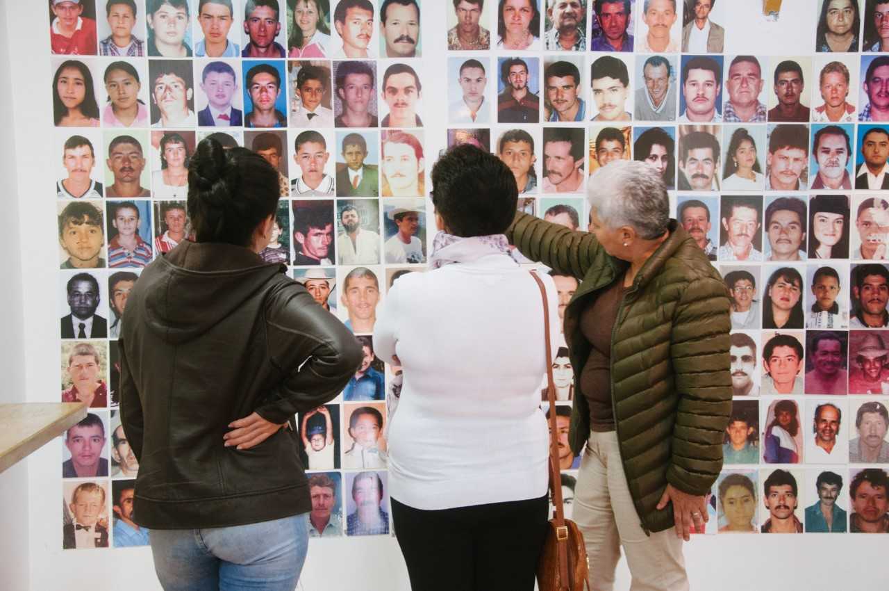 Tres mujeres mayores están de espaldas a la cámara, de frente a una pared llena de fotos de personas que fueron desaparecidas.