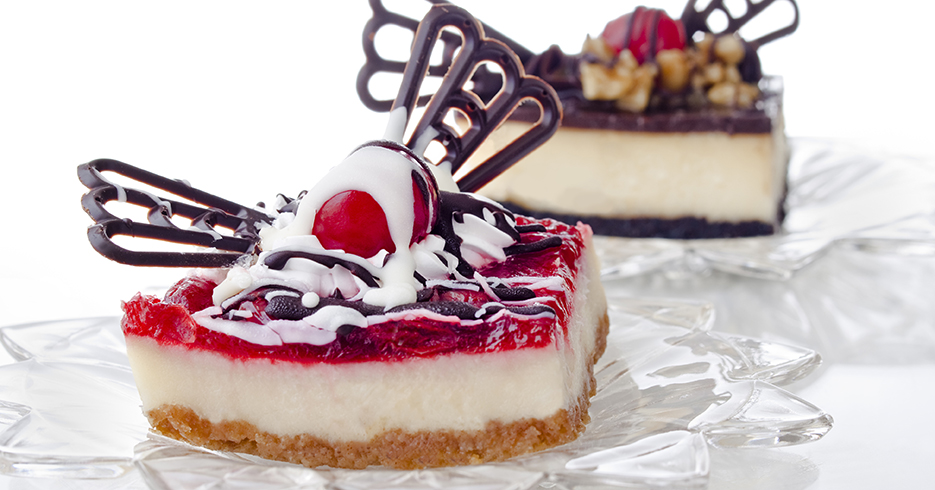 A strawberry cheesecake in the foreground with a chocolate cheesecake in the background. The focus is on the cherry.