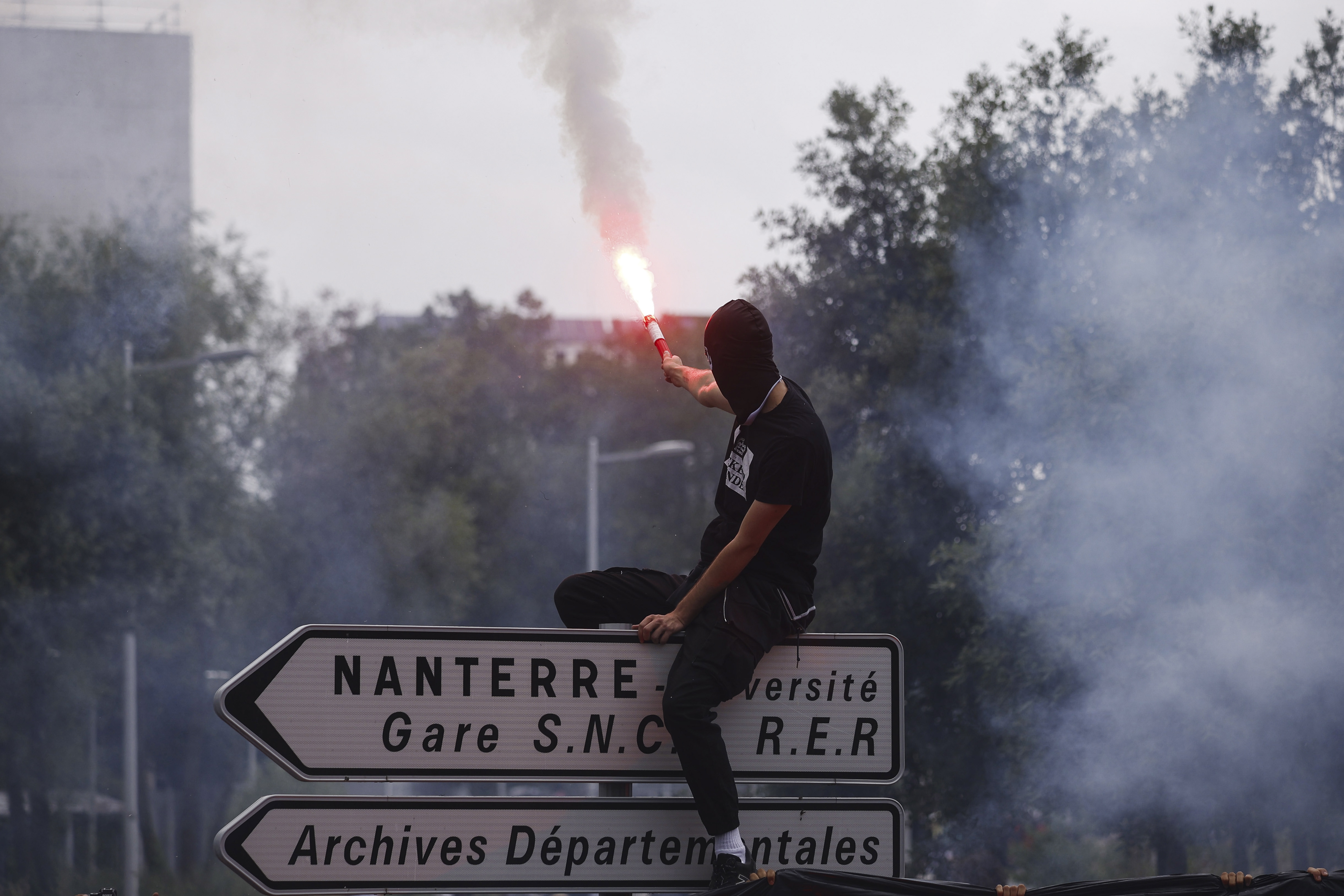 Nanterre (France), 29/06/2023.- A black-clad protester, perched atop a traffic sign, holds a flare during a march in the memory of 17-year-old Nahel, who was killed by French Police in Nanterre, near Paris, France, 29 June 2023. Violence broke out after the police fatally shot a 17-year-old during a traffic stop in Nanterre on 27 June. According to the French interior minister, 31 people were arrested with 2,000 officers being deployed to prevent further violence. (Protestas, Disturbios, Francia) EFE/EPA/YOAN VALAT