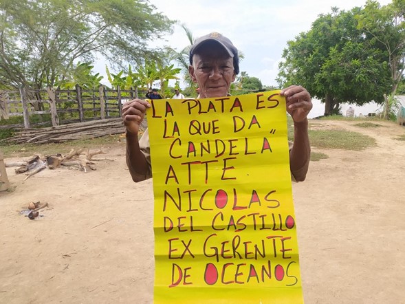 Un señor de cachicha muestra una pancarta de color amarillo con un mensaje: "La plata es la que da candela"