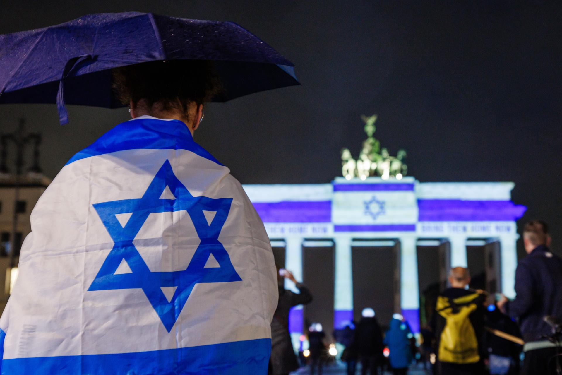 Berlin (Germany), 07/10/2025.- A person with an umbrella and a flag of Israel around the shoulders stands in front of the Brandenburg Gate that reads 'Bring them home now' and shows the Israeli flag during an illumination of the Brandenburg Gate to mark the second anniversary of the Hamas attack against Israel in Berlin, Germany, 07 October 2025. (Alemania) EFE/EPA/CLEMENS BILAN