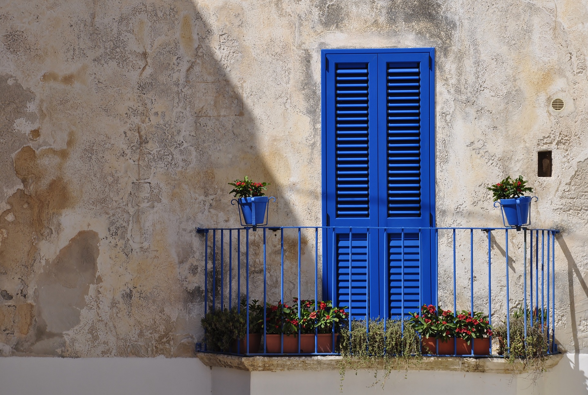 Foto que muestra un pequeño balcón de rejas metálicas con una puerta azul de madera en el que hay plantas sembradas en materas.