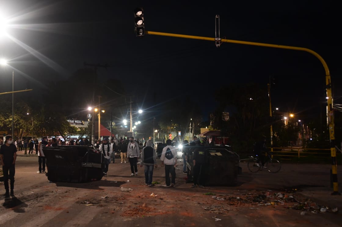 Dos jóvenes de espaldas junto a dos contenedores de basura que fueron puestos en medio de la vía.