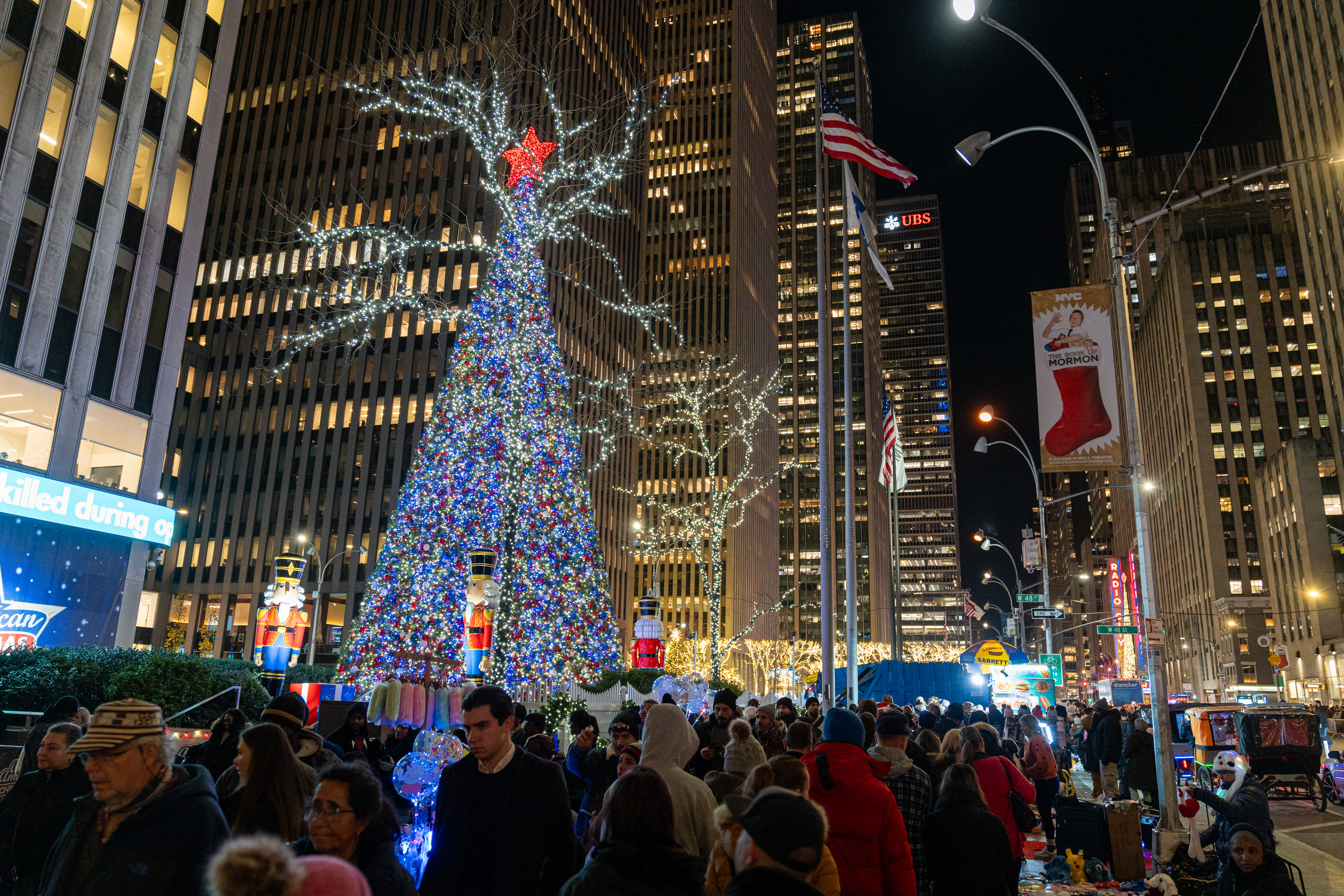 ACOMPAÑA CRÓNICA: EEUU NAVIDAD - AME5017. NUEVA YORK (ESTADOS UNIDOS), 18/12/2023.- Personas caminan junto a la decoración navideña y el árbol de Navidad en la Sexta Avenida en Fox Corner, el15 diciembre de 2023, en Nueva York (EE.UU.). La Navidad en Nueva York, como en el resto de las grandes metrópolis del mundo, ha perdido casi por completo su original sentido religioso y se reduce a poco más que un pino decorado con figuritas intrascendentes y luces, así como coronas vegetales, aunque en esta ciudad la corrección política llega a extremos un tanto peculiares. El tradicional saludo de 'Feliz Navidad' ('Merry Christmas') ya cayó en desuso: en esta ciudad poblada por gentes de todas las razas y religiones se ha optado hace tiempo por el más neutral 'Felices fiestas', y las escuelas ya no dan 'vacaciones de Navidad', sino 'vacaciones de invierno'. EFE/ Angel Colmenares