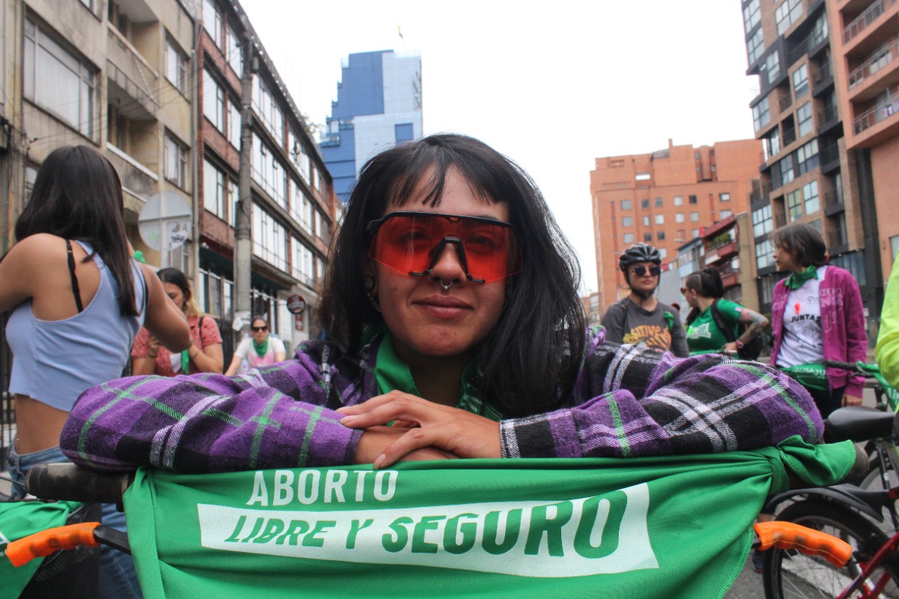 Una mujer con gafas recostada sobre el manubrio de una bicicleta en la que hay una pañoleta de aborto libre