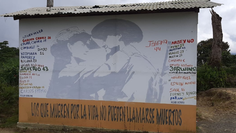 Una pared con con fondo blanco, tiene a sus laterales los nombres de personas excombatientes asesinadas.