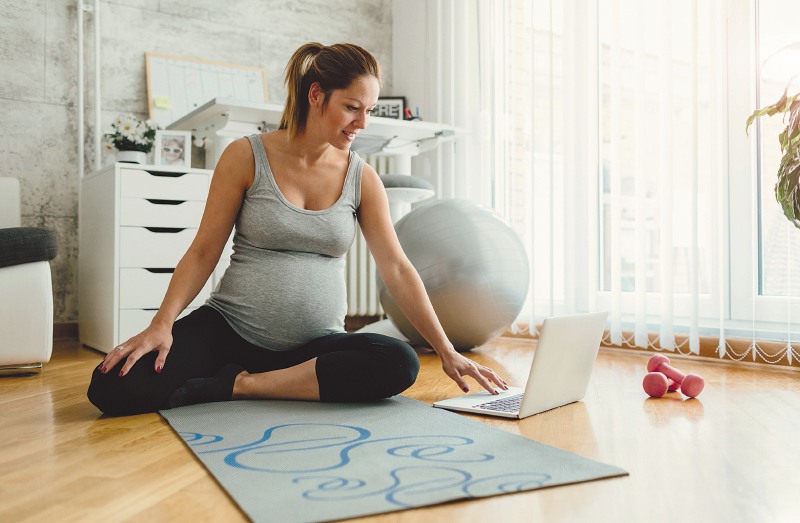30 weeks pregnant woman exercising at home. Sitting on exercise mat, making a break and using laptop. Looking for exercises on the internet.