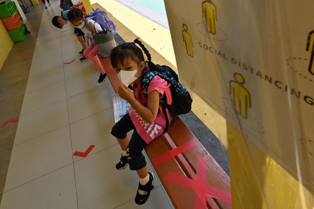 Estudiantes de Escuela Internacional de Pioneros en Bangkok esperan sentados en una banca que tiene marcas para garantizar el distanciamiento social.