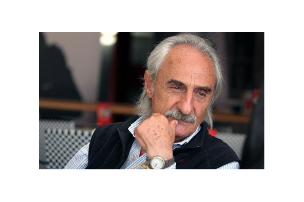 Alfredo Molano Bravo está vestido con una camisa azul clara con rayas blancas y tiene encima un chaleco. Él tiene su mano izquierda apoyada sobre su cara y no mira a la cámara.